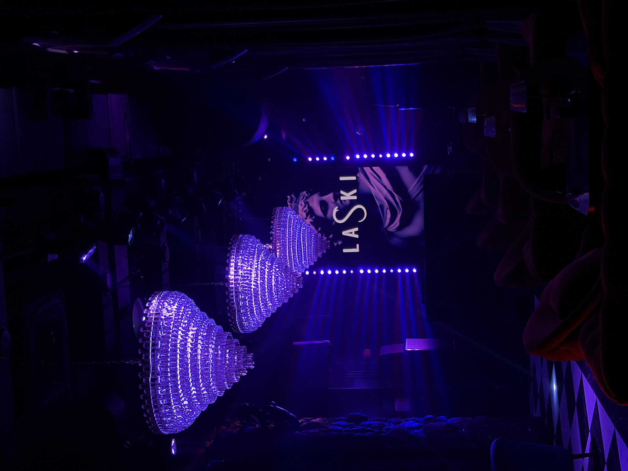 This image depicts the interior of Laski Bar, located in Moscow, Russia. The scene is illuminated primarily by intense purple lighting, creating a dark, opulent atmosphere. Three large, tiered crystal chandeliers hang from the dark ceiling, glowing with purple light. On the back wall, a large screen or projection displays the stylized white text "LASKI" overlaid on an image of a classical male bust sculpture, also bathed in purple light. Vertical strips of bright purple LED lights flank this central display, emitting horizontal light beams across the room. In the foreground and to the right, multiple dark red upholstered lounge chairs and sofas are arranged, largely silhouetted, with small, dimly lit tables featuring reflective-shaded lamps interspersed among them. On the left side, a section of a black and white checkered floor is visible, reflecting the purple light, next to a darkly textured wall resembling rock or rough stone. No individuals are present in the scene.