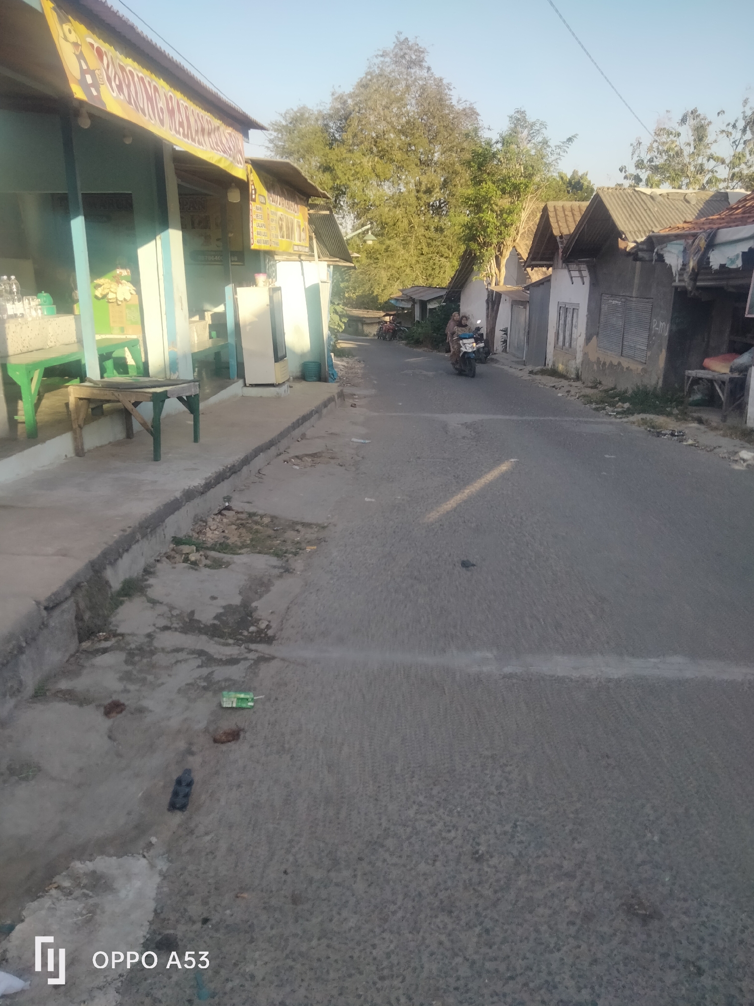 The image depicts a street scene in what appears to be a village or a less urbanized area, likely in Badung, Indonesia, given the visual cues of the architecture and vegetation. The photograph is taken during daylight, with the sun casting long shadows.

On the left side of the street, there is a building with a green and blue facade, possibly a shop or a small eatery, indicated by a yellow banner with Indonesian text that partially reads "WARUNG MAKAN". Inside, several bottles of water and what appear to be packaged snacks are visible on a counter. Green benches and a wooden table with green legs are placed on the sidewalk in front of the establishment. A white refrigerator stands near the entrance.

The street itself is paved with asphalt and appears to be two lanes wide. A white line marks the center of the road. On the right side of the street, there are several small houses with pitched roofs, some made of corrugated metal. The architecture is modest and suggests a residential area. Vegetation, including trees with dense foliage, is visible behind the houses and on the left side of the street.

A motorcycle is seen on the right side of the road, traveling away from the viewer. Two individuals are riding on the motorcycle; one is driving, and the other is seated behind. They are both wearing traditional Indonesian clothing.

The ground on the left side of the street, adjacent to the sidewalk, is a concrete drainage channel with some debris and vegetation growing within it. The overall environment appears somewhat weathered, with signs of age and possibly some disrepair in the infrastructure. The sky is clear and a light blue, indicating good weather. The bottom of the image includes a watermark with the text "OPPO A53".
