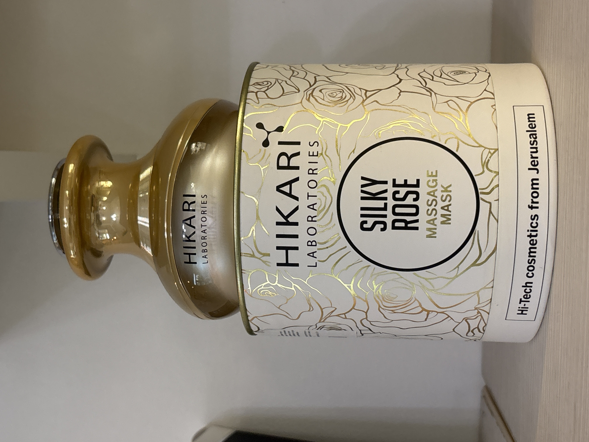 The image shows a cosmetic product from "HIKARI LABORATORIES," specifically a "SILKY ROSE MASSAGE MASK," described as "Hi-Tech cosmetics from Jerusalem." The product packaging consists of a cylindrical container with a lid. The container has a white background with a gold rose pattern. The brand name "HIKARI LABORATORIES" is printed in black letters, with a logo above. A black circle surrounds the words "SILKY ROSE" and "MASSAGE MASK" printed in white. The lid appears to be made of gold colored plastic. The image was taken indoors. The location in the city of Vsevolozhsk, Russia is not apparent from the image.