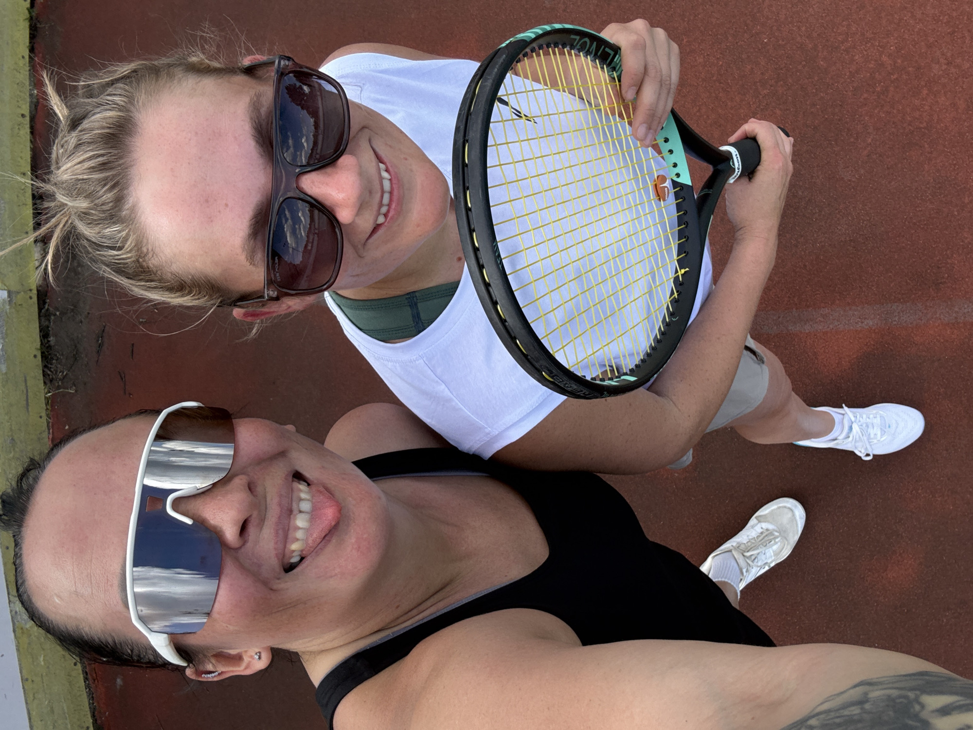 This media file features two women on a tennis court, engaged in what appears to be a casual game or practice session. The perspective is a high-angle, looking down at the subjects.
The woman in the foreground is wearing a black tank top and large, white-framed sunglasses that reflect the sky. She has dark hair pulled back and is smiling broadly, with her tongue slightly out. The woman behind her is wearing a white tank top and dark sunglasses, with her blonde hair tied up. She is holding a tennis racket, suggesting they are about to play or have just finished. She is also smiling.
The ground is a reddish-brown tennis court surface. Both women are wearing athletic shoes. The overall mood seems lighthearted and energetic, indicative of a fun outdoor activity. The lighting suggests it is daytime, likely sunny, given the reflections in the sunglasses. No specific weather conditions are evident beyond the sunshine.
There are no discernible buildings or geographical landmarks that provide further location context, other than the tennis court itself. The provided location context is "Chaikovsky, Russia." There is no visible text in the image.