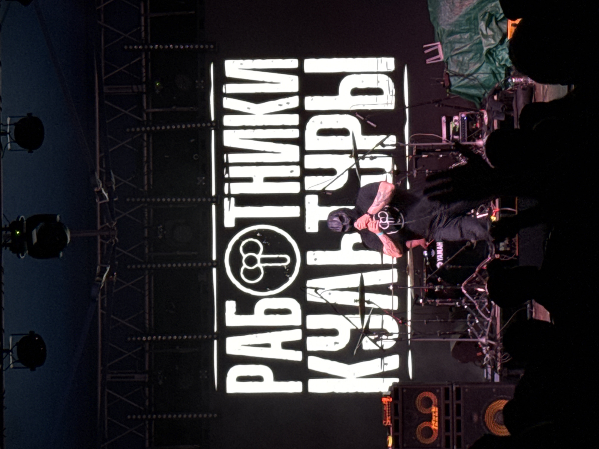 The image depicts an indoor live music performance in Большая Кивара, Russia. Centered on the stage, a performer wearing a dark mask and a dark t-shirt with a distinct circular logo is holding a microphone, actively engaging in the performance. The logo features a central vertical line with two symmetrical loops at its top. Behind the performer, a large illuminated screen displays the Cyrillic text "РАБОТНИКИ КУЛЬТУРЫ" (Workers of Culture) in bold white letters, with the aforementioned circular logo incorporated into the first word.
Stage equipment visible includes a drum kit with a "YAMAHA" branded kick drum behind the performer, and two stacked speaker cabinets with illuminated circular patterns on their grilles positioned to the viewer's left. Various microphone stands, cables, and electronic devices are scattered across the stage. To the viewer's right, a green tarp covers stage equipment, with a white, U-shaped object resting on it. Overhead, metal truss structures support dark stage lights and other rigging. In the immediate foreground, a crowd of audience members is silhouetted, with some hands raised towards the stage. The overall lighting is dim, emphasizing the illuminated screen and the performer.