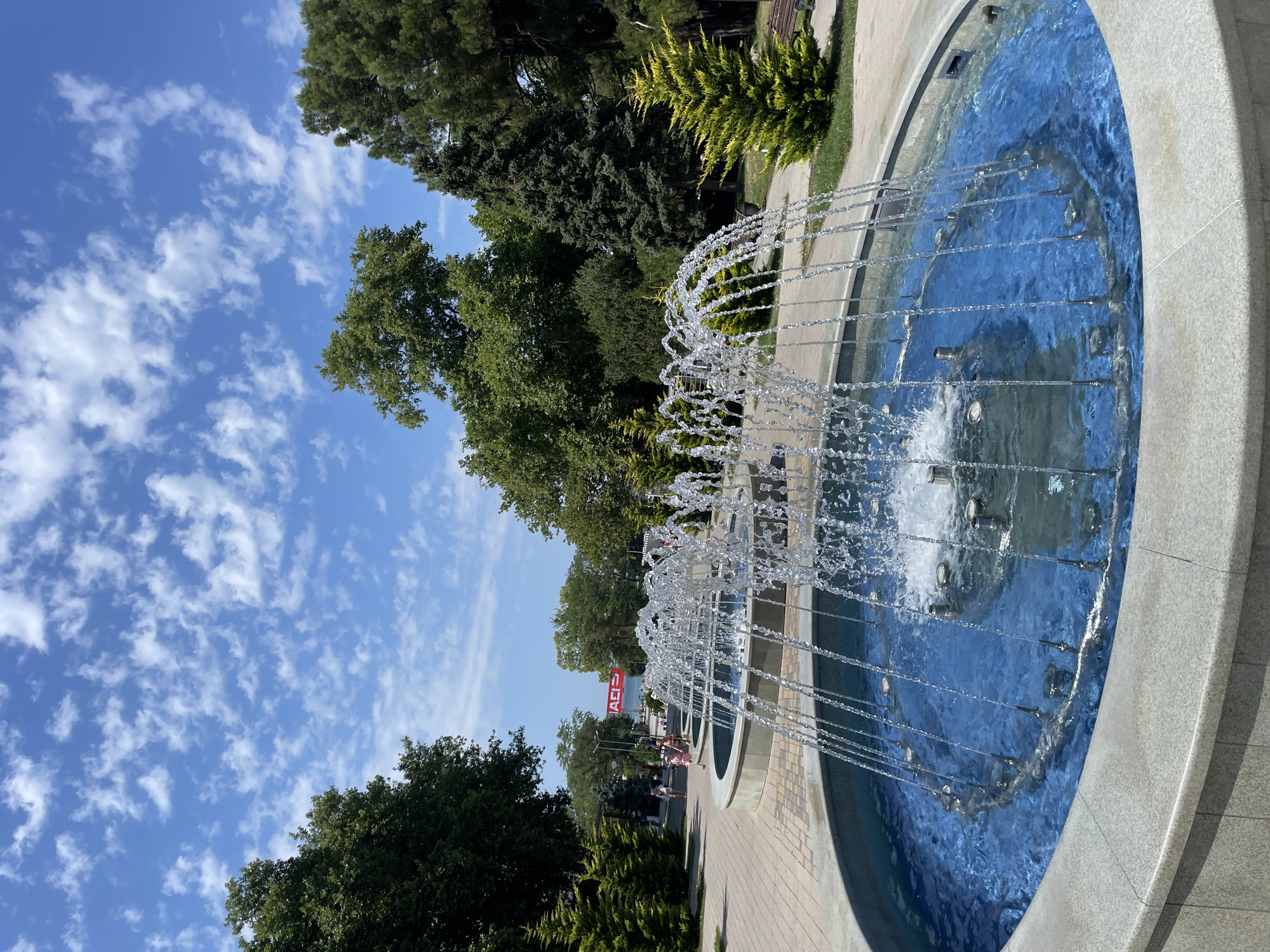 The image depicts a bright, sunny day in Anapa, Russia, with a foreground focus on a multi-tiered fountain with water spraying upwards. The fountain basin is circular and filled with vibrant blue water. Beyond the fountain, a paved walkway meanders through a park-like setting. Lush green trees, including various conifer species, line the walkway, creating a shaded canopy. Several people, including adults and children, are visible strolling along the path in the mid-ground. In the distance, a large red sign with white lettering is visible, suggesting a commercial area or attraction. The sky above is a clear blue, dotted with scattered white clouds.