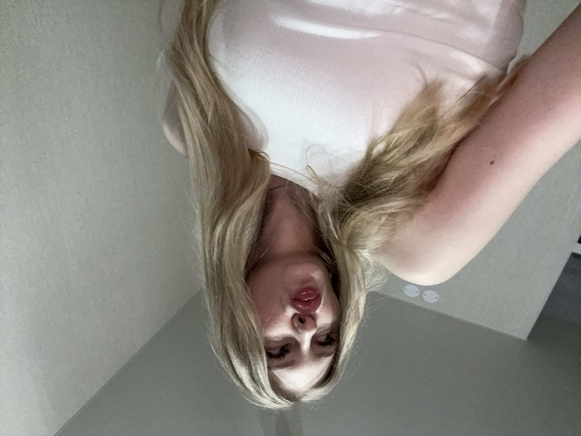 The media file presents an indoor portrait of a person with long, blonde hair, captured and displayed in an inverted orientation. The subject's head is positioned towards the bottom of the frame, with their body extending upwards. They are wearing a white, ribbed tank top or camisole. Their blonde hair flows around their face and shoulders.
The person's face is directed towards the viewer, with their lips pursed in a pronounced, glossy red or pink pout. Their eyes are partially visible, adorned with dark eyelashes. A fair-skinned arm is visible on the right side of the frame, featuring a small dark mole or freckle.
The background primarily consists of plain, light grey walls, indicating an indoor setting. A subtle corner where two walls meet can be seen towards what would be the top right if the image were correctly oriented. In the bottom right section (which would be the top right when rotated), two small, circular, light-colored fixtures or markings are visible on a darker grey surface, likely a wall or ceiling.
The image suggests a playful selfie, possibly taken from a low angle. There is no visible text. The scene is located indoors, and the location context provided is Perm, Russia.