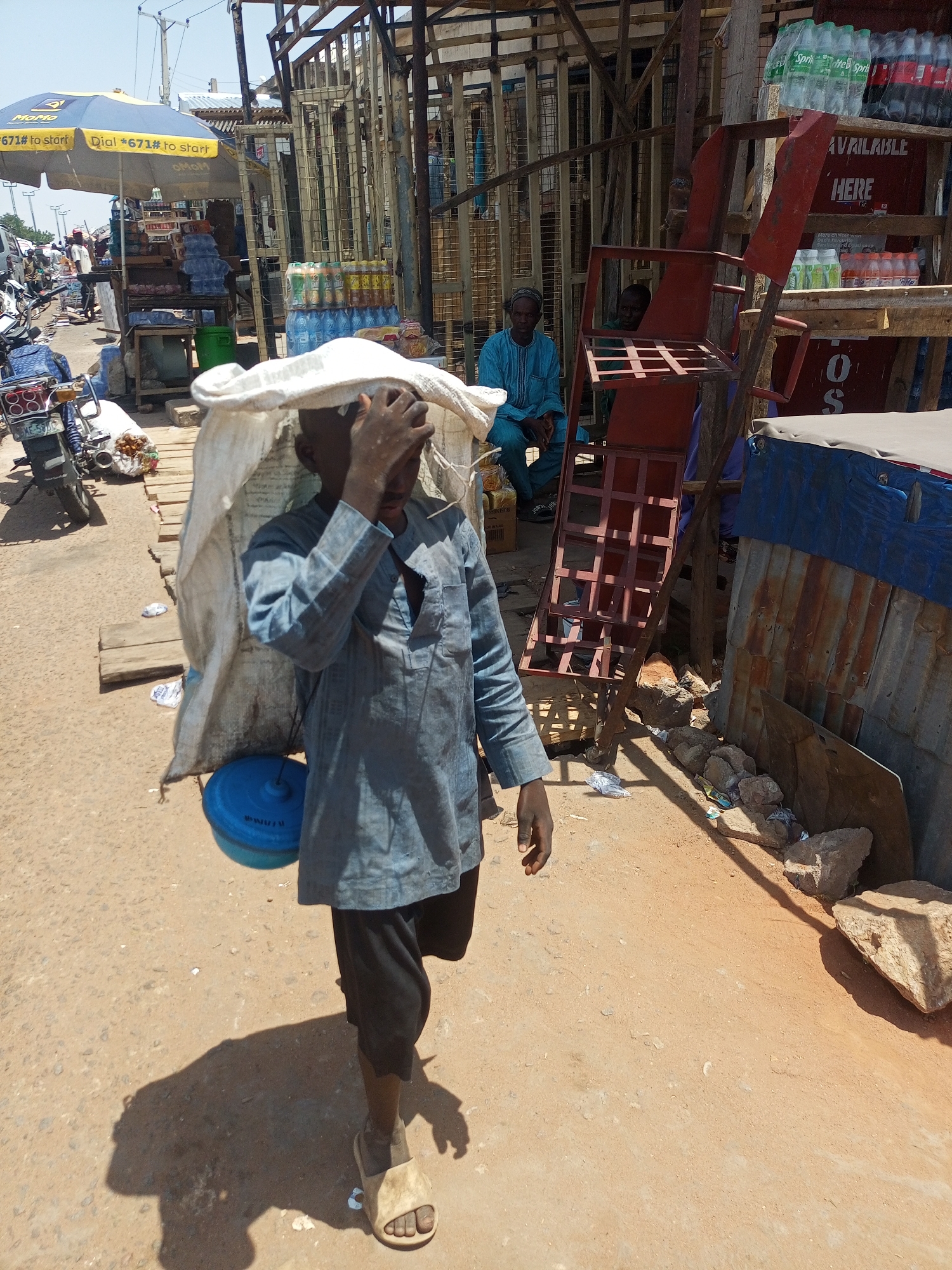 This medium shot captures a bustling market scene in Gombe, Nigeria, under bright mid-day sun, evident from the strong shadows cast on the dusty ground. The primary subject, a young person, walks towards the viewer, carrying a large, heavy-looking white sack on their back/head. They wear a light blue, long-sleeved tunic over dark trousers and light-colored sandals. One hand is raised, shielding their eyes from the sun, while a blue plastic container hangs from their left side.
Behind the foreground subject, various market stalls are visible. To the left, a "MoMo" branded blue and yellow umbrella provides shade for a vendor, with stacks of bottled water and other beverages displayed on wooden tables. A motorcycle is parked nearby. In the background, two men are seated in the shade; one wears a blue traditional outfit.
To the right, a stall features shelves displaying more bottled drinks, including Sprite and Coca-Cola, with a sign reading "AVAILABLE HERE." Further right, a makeshift structure of corrugated metal sheets and wooden planks forms part of a stall. The ground is unpaved, covered in dirt and sand, with scattered rocks and some litter.
Visible text includes "MoMo" and "Dial *671# to start" on the umbrella, "Sprite" on drink bottles, and "AVAILABLE HERE" on a red shelf structure. The overall scene depicts daily commerce and life in an outdoor market environment.