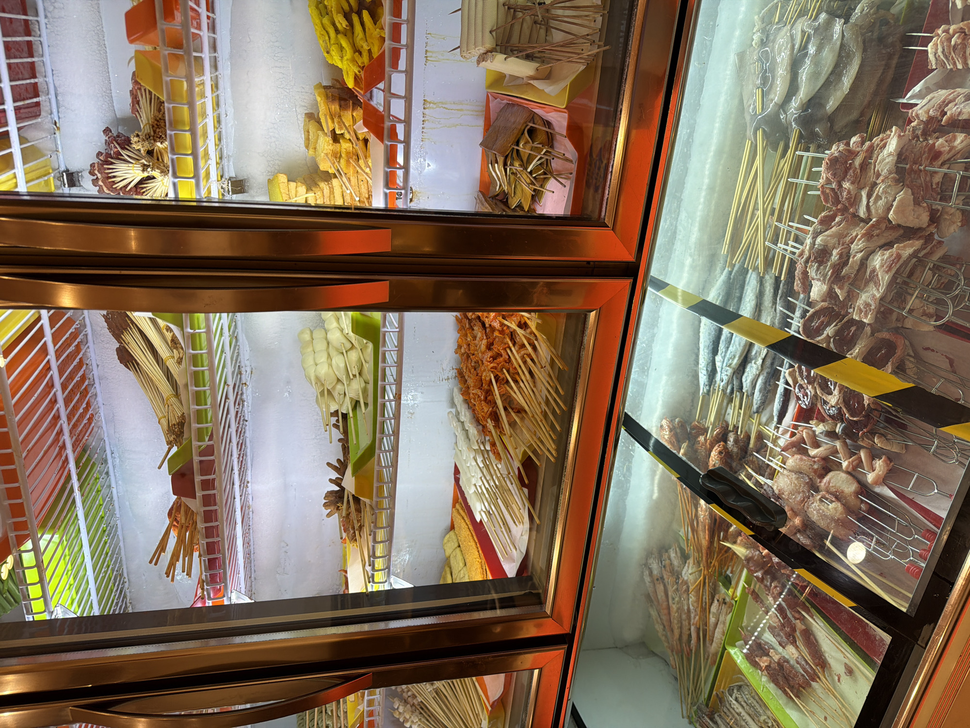 A refrigerated display case in what appears to be a food establishment in Chongqing High-tech Zone, China, contains various skewers of food. The skewers include meat, seafood, vegetables, and other processed food items, all lined up neatly on shelves. The refrigeration unit is well-lit, with frost visible on the interior walls and shelves. The scene is viewed from a high angle, capturing the contents of the display case.