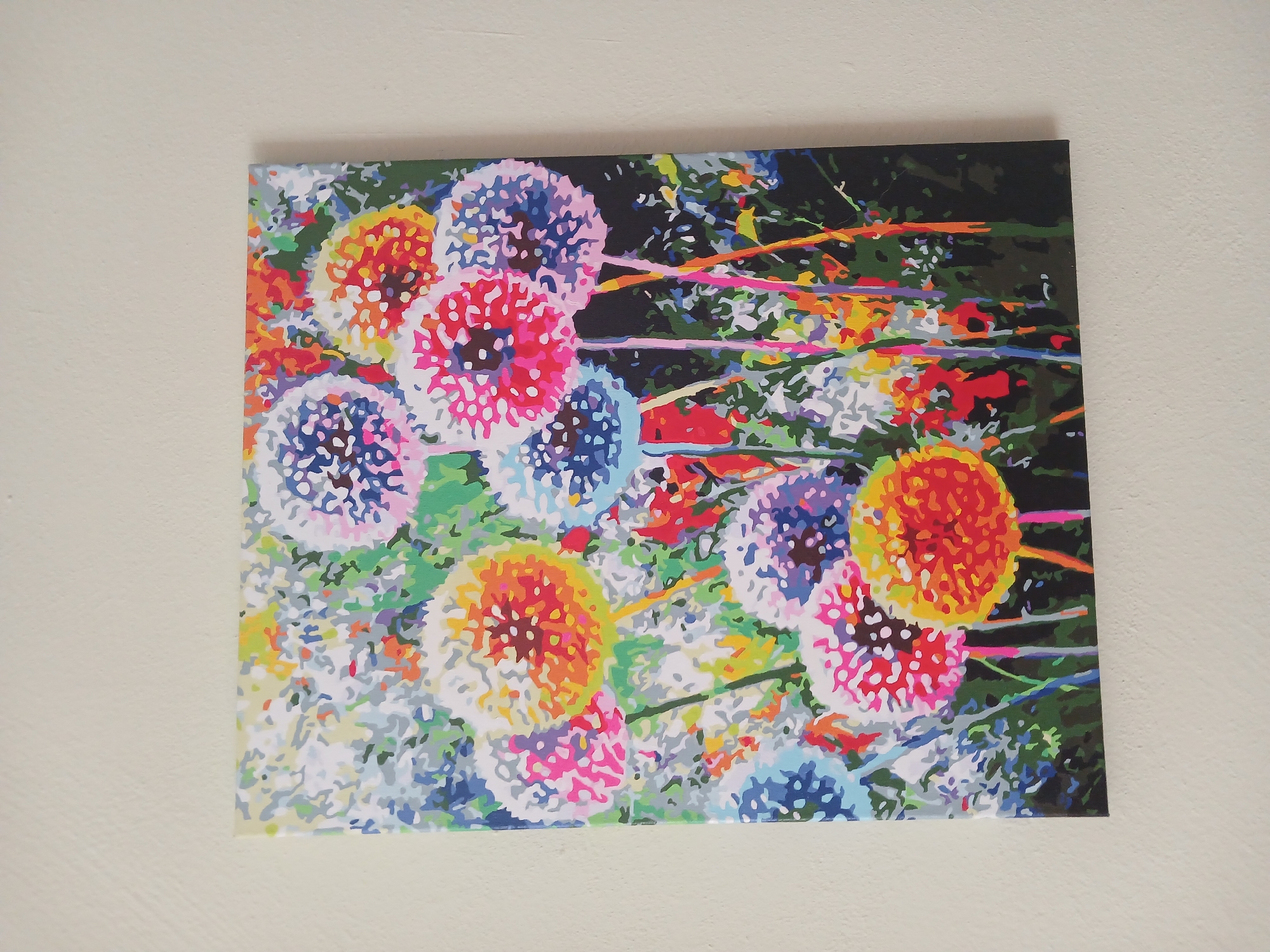 A brightly colored painting of dandelions hangs on a light-colored textured wall. The painting depicts a cluster of dandelions in various stages of bloom, rendered in bold, abstract brushstrokes of pink, orange, yellow, blue, and white. The dandelions are set against a backdrop of green foliage and hints of other small flowers. The painting is presented in a landscape orientation. The location is Mamaivtsi, Ukraine.