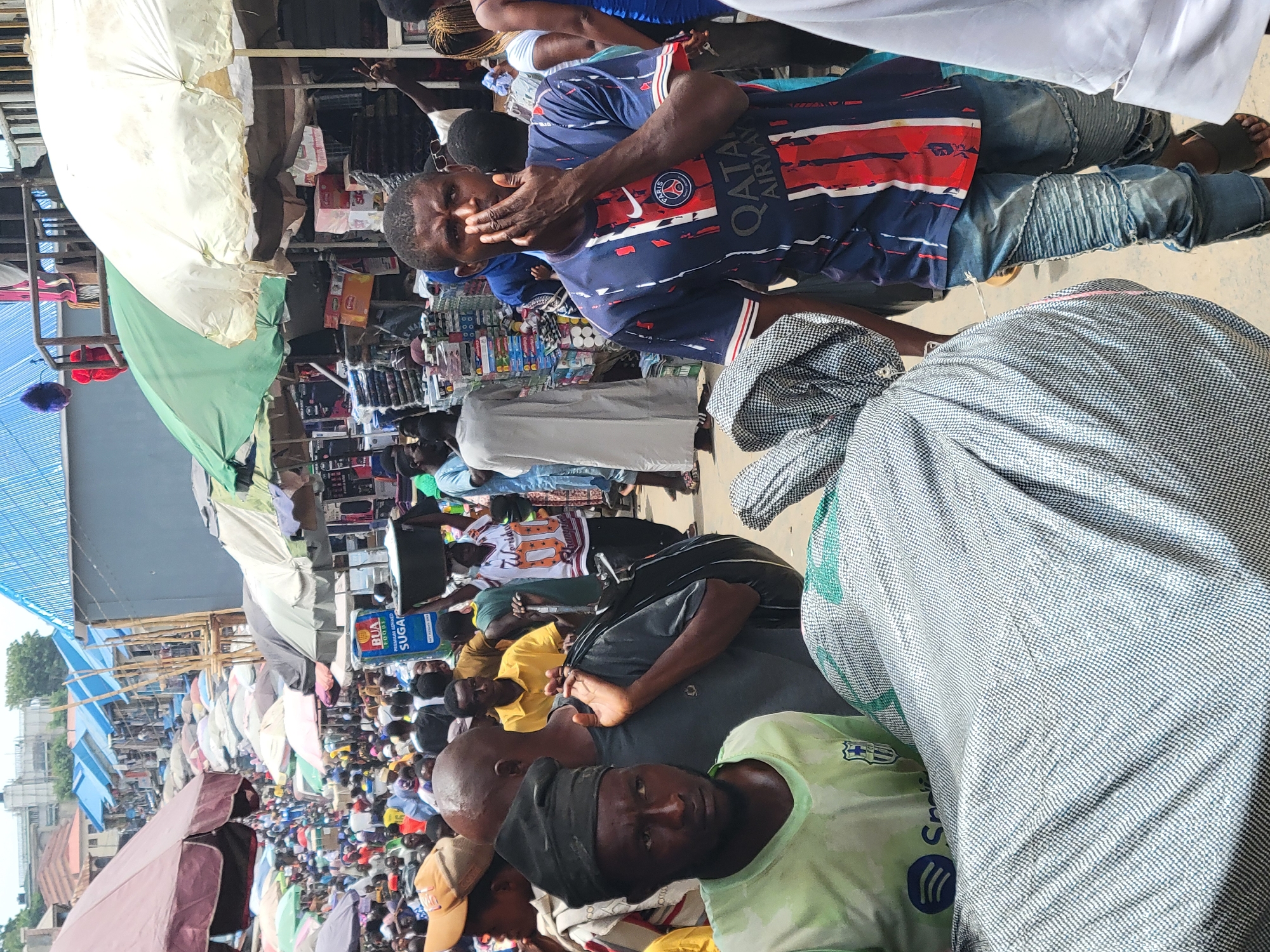 The image captures a crowded marketplace scene in Suleja, Nigeria. People are densely packed, and many vendors have set up stalls with goods covered by umbrellas for shade. One man wears a blue and red soccer jersey, holding his hand near his face. There are various goods on display at the stalls, including boxes of sugar. Many people are looking directly at the camera.