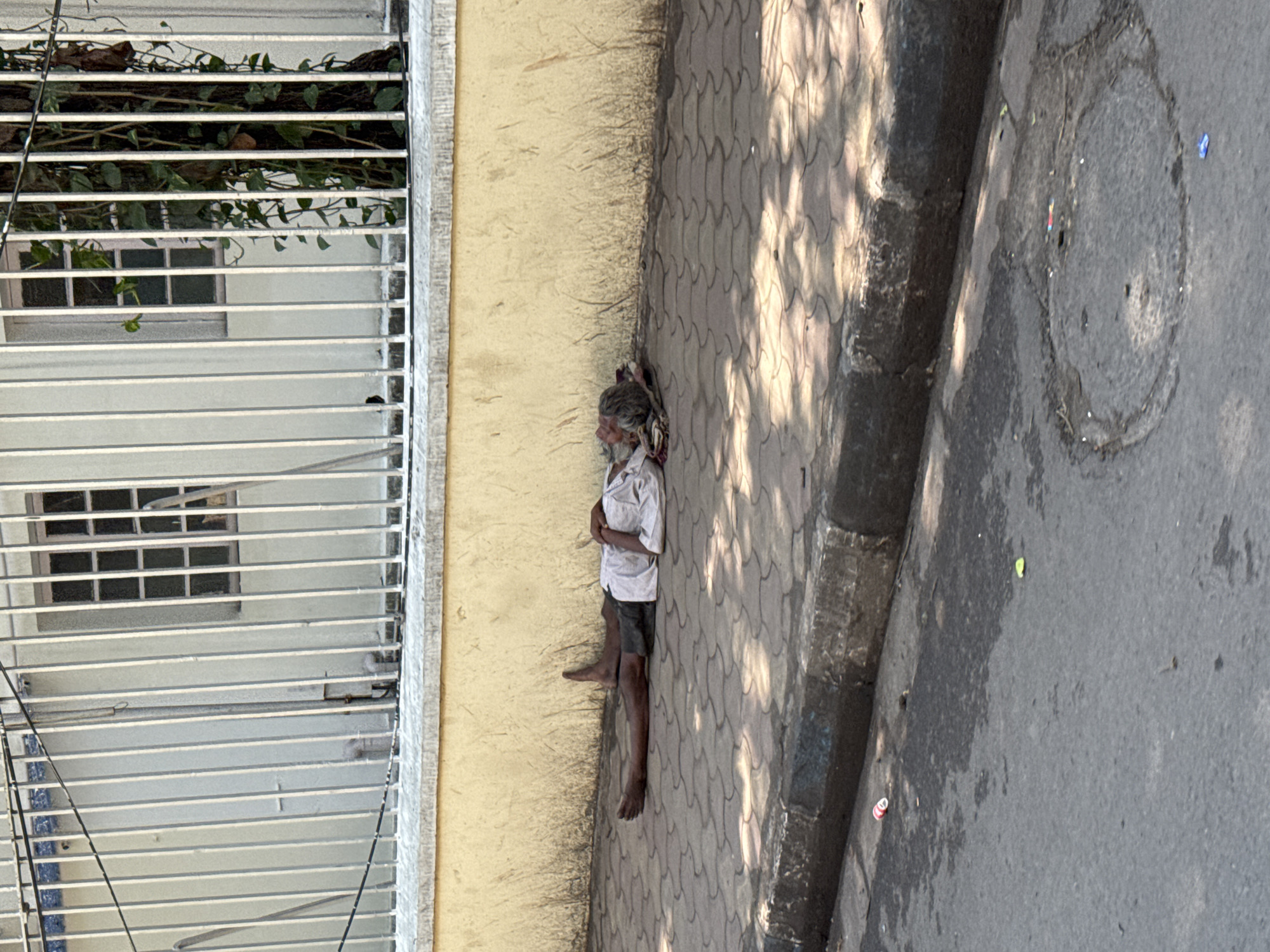 The image captures an outdoor scene, likely in an urban or semi-urban environment, with a focus on an individual resting on a sidewalk next to a building. The image is oriented vertically, showing the scene from an elevated perspective looking down.
In the center of the frame, an older individual, presumably male, is seen lying on their side against a light yellow wall. The person has long, tangled, grey and white hair and a matching beard. They are barefoot and wearing a light-colored, short-sleeved shirt and dark shorts. Their posture suggests they are resting or sleeping, with their eyes closed or downcast, and a bundle of fabric serves as a makeshift pillow under their head.
To the right of the person is a sidewalk made of tan-colored interlocking paver blocks, which transition into a dark grey curb, and then an asphalt road. The sidewalk and parts of the road are covered in dappled shadows, indicating bright sunlight from an unseen source, suggesting it is daytime. Small pieces of litter, including blue and red fragments, are visible on the asphalt road, where a circular manhole cover is also embedded.
To the left of the person is the base of the yellow wall, which has a distinct line of dark, streaky discoloration or dirt running horizontally along its lower section. Above this wall, a building structure is visible, featuring multiple horizontal white metal grates or louvers. Behind these grates, parts of a window with several panes can be seen. Green vines and foliage are growing through and over the top sections of these grates, adding a touch of natural growth to the otherwise structured environment. No discernible text is visible in the image.