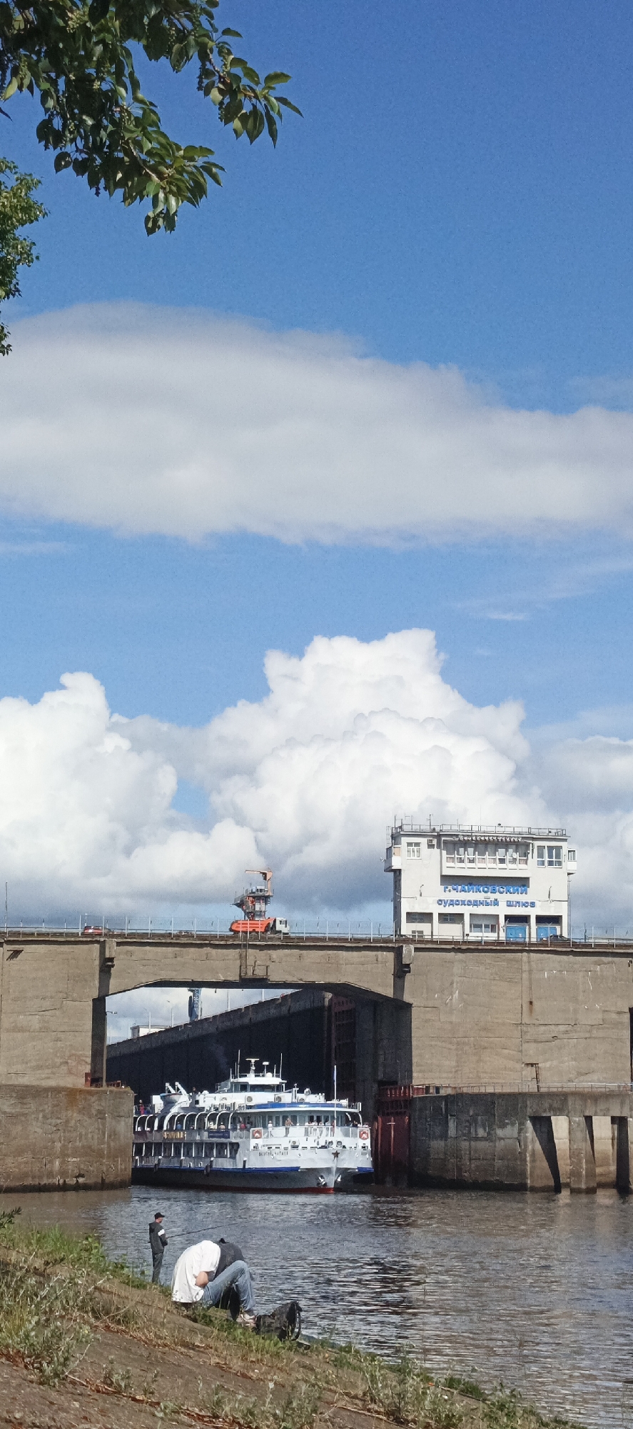 The image shows a river lock with a passenger ship inside. The lock's control tower is visible on the right side of the bridge above the lock. In front of the lock, on the riverbank, two individuals are present, one seated and another standing nearby. The lock is identified as "Судоходный Шлюз" in Russian, which translates to "Navigation Lock," and is associated with the city of Chaikovsky (Г. Чайковский) in Russia. The sky is predominantly blue with large, puffy white clouds. A few green tree branches are visible in the upper left corner of the frame. An orange construction crane is also present on top of the bridge.