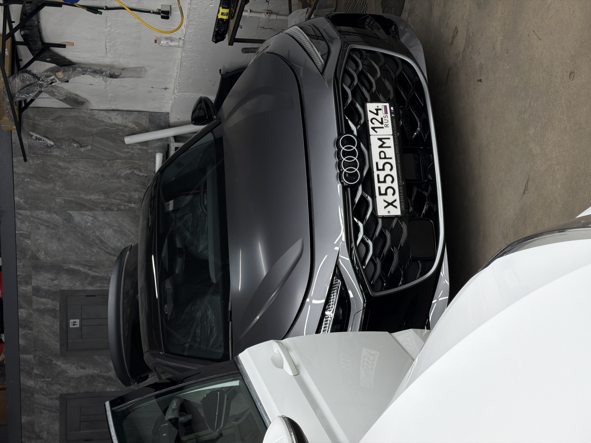The image captures a close-up view of two cars parked indoors. The primary focus is on the front of a grey Audi, specifically its grille and the Russian license plate "X555PM 124 RUS". The license plate also contains smaller text mentioning "Элит Авто Сибирь," which is an official BMW dealer. This detail suggests that the Audi could be parked at or near the mentioned car dealership in Krasnoyarsk, Russia, based on the region code on the license plate. Adjacent to the Audi is a white car, partially visible. The background shows sections of walls, one with a marble-like pattern and another with a set of gray, electrical or utility boxes. Other details such as a table and hanging cables are visible in the background as well.