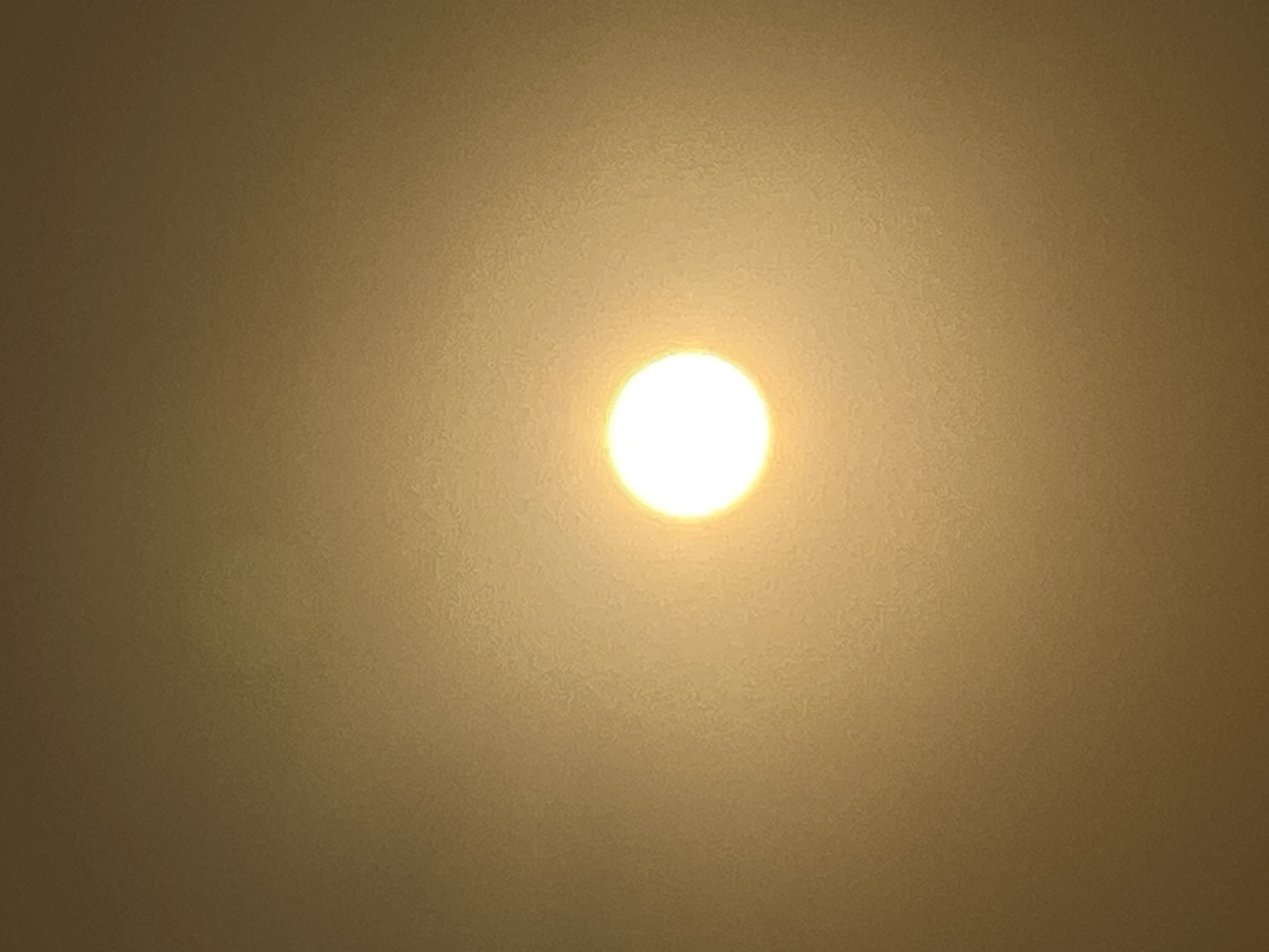 A bright, round orb of light, likely the sun, is centrally located in the image. The background is a hazy, muted yellow or golden color, suggesting an atmospheric condition like fog, smog, or an overcast sky. There are no discernible people, subjects, objects, actions, or interactions visible in the image. The scene is abstract and lacks specific environmental context within Kuta, Indonesia, or any other city.
