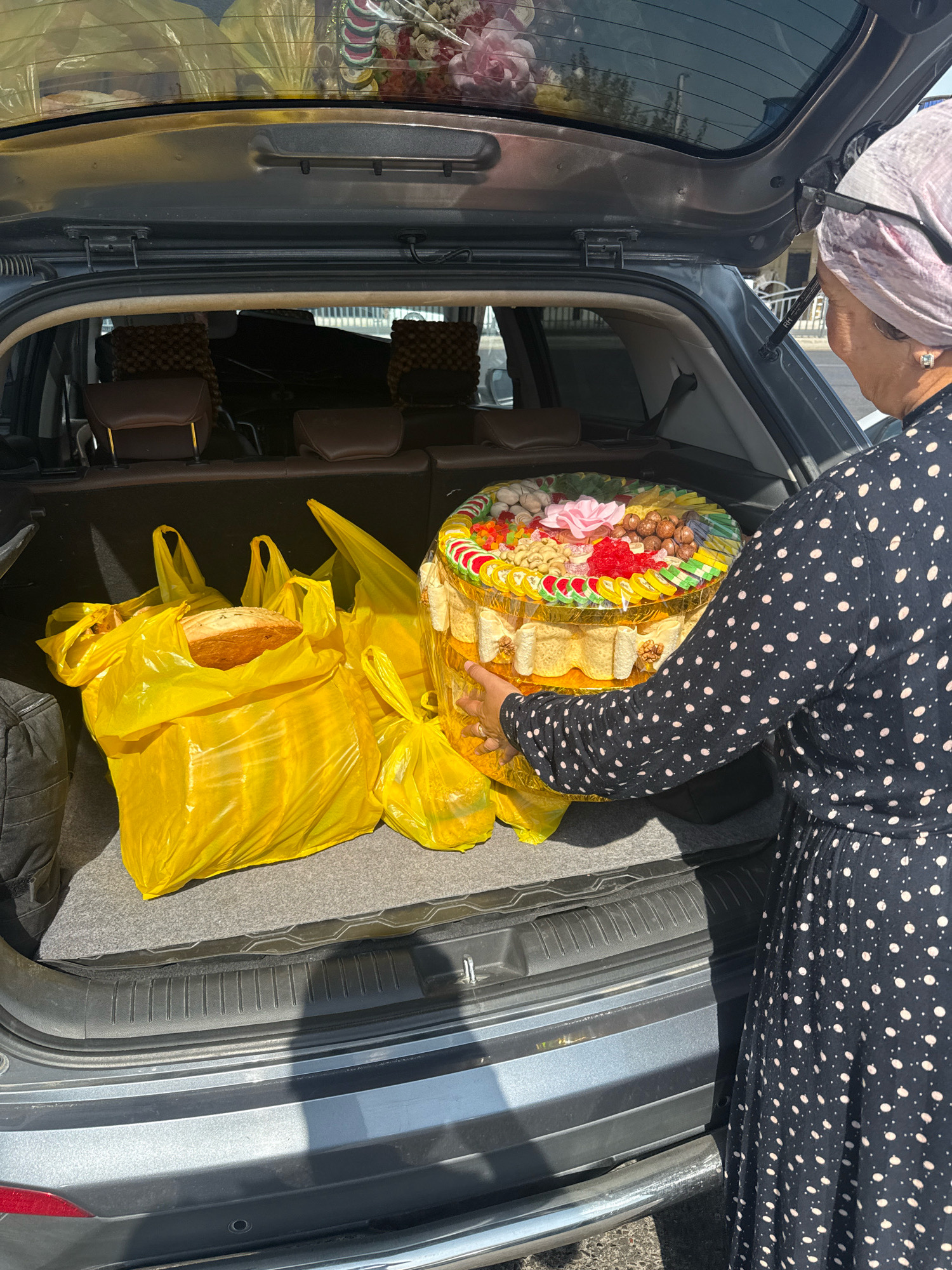 An individual, wearing a black dress with white polka dots and a light pink headscarf, is placing a large, round, tiered tray of nuts, dried fruits, and candies into the open trunk of a gray SUV. The tray is decorated with a pink flower on top. To the left of the tray are three large, bright yellow plastic bags, one containing a loaf of bread. The interior of the trunk is lined with dark gray carpeting. The vehicle's rear seats are visible behind the items in the trunk. The scene appears to be outdoors, possibly on a street, with glimpses of buildings and foliage in the background, suggesting a location in Tashkent, Uzbekistan. The lighting indicates it is daytime.