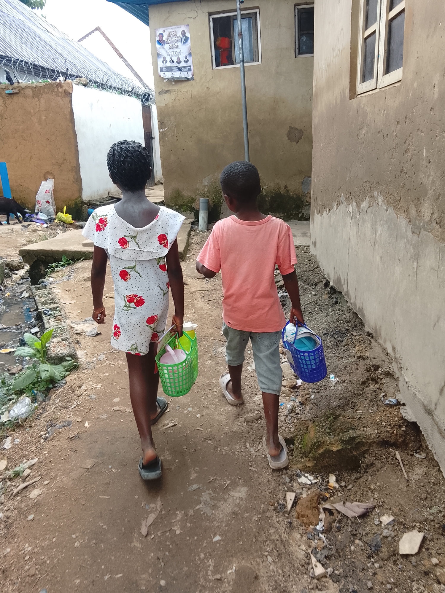 The image shows two young children walking away from the camera down a dirt path in Abuja, Nigeria. The child on the left, facing away, is wearing a white dress with red floral patterns and carrying a green plastic basket containing a pink bucket and other items. The child on the right is wearing a pink t-shirt and denim shorts and is carrying a blue plastic basket. The path is lined with dilapidated buildings on both sides. The building on the left has a corrugated metal roof and barbed wire along the top of a wall. A poster with text and images of people is affixed to the wall of the building on the right. The ground is littered with debris, including plastic bags, leaves, and what appears to be sewage in a ditch to the left of the path. The overall environment suggests a low-income, urban setting.