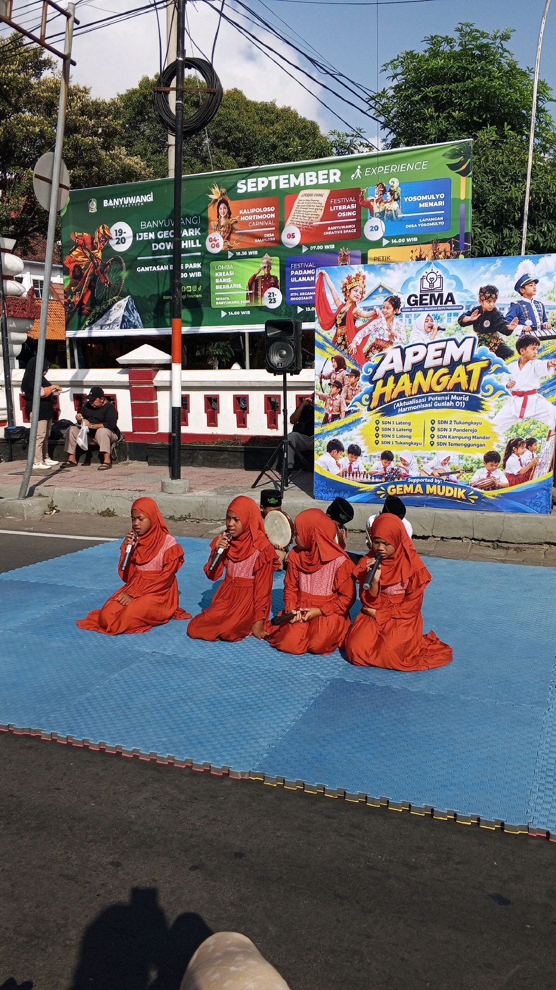 A group of young children, dressed in orange Islamic dresses and headscarves, are performing on a blue mat placed on an asphalt road in Tamanbaru, Indonesia. They are seated and holding microphones, suggesting a singing or speech performance. One child in the background is playing a drum. Behind the children, a large banner displays various events and activities, including "BANYUWANGI SEPTEMBER EXPERIENCE" and advertisements for different festivals and programs, indicating a community event or festival. The environment includes a street, sidewalk with people sitting, a lamppost, and trees in the background, all under a bright sky.