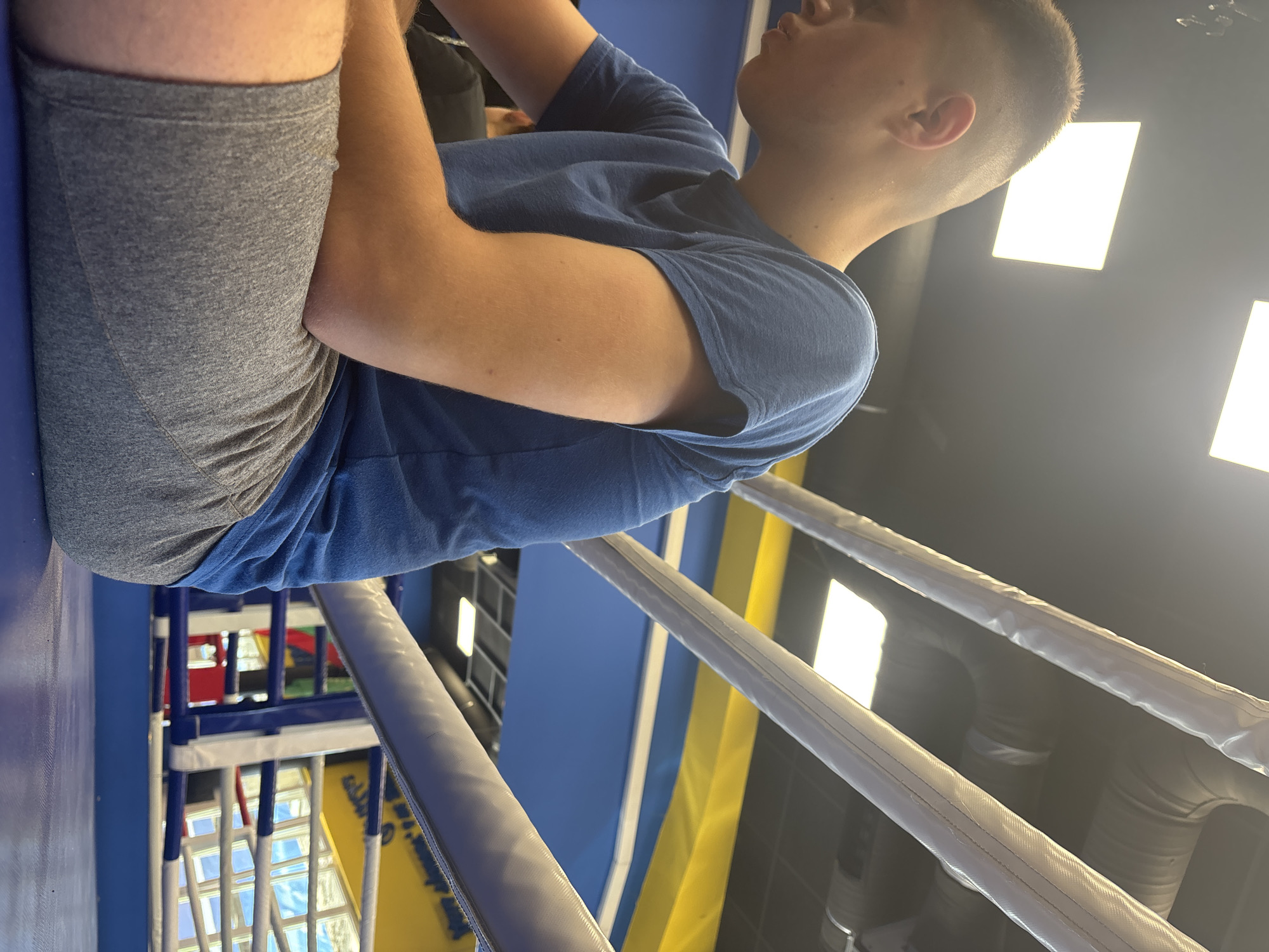 A young male is depicted sitting on the edge of a boxing ring, viewed from a low, slightly upward and side angle. He has short hair and is wearing a blue t-shirt and grey athletic shorts. His face is mostly obscured as he looks upwards and towards the right of the frame. He appears to be resting or observing, with his arms positioned near his lap.
The setting is an indoor athletic facility, specifically a boxing gym or a similar training center in Sofia, Bulgaria. The foreground features the white, padded ropes of a boxing ring. Beyond the ring, a blue and yellow matting or wall section is visible, contributing to the vibrant color scheme of the facility. The ceiling is dark and features multiple bright, rectangular overhead lights, as well as visible ventilation ducts, suggesting an industrial or modern gym design. In the background, behind the ropes, there are glimpses of other gym equipment or structures, characterized by blue, white, and red bars, possibly part of a multi-sport area. No specific time of day can be determined due to the indoor, artificially lit environment. There is no visible text in the image.