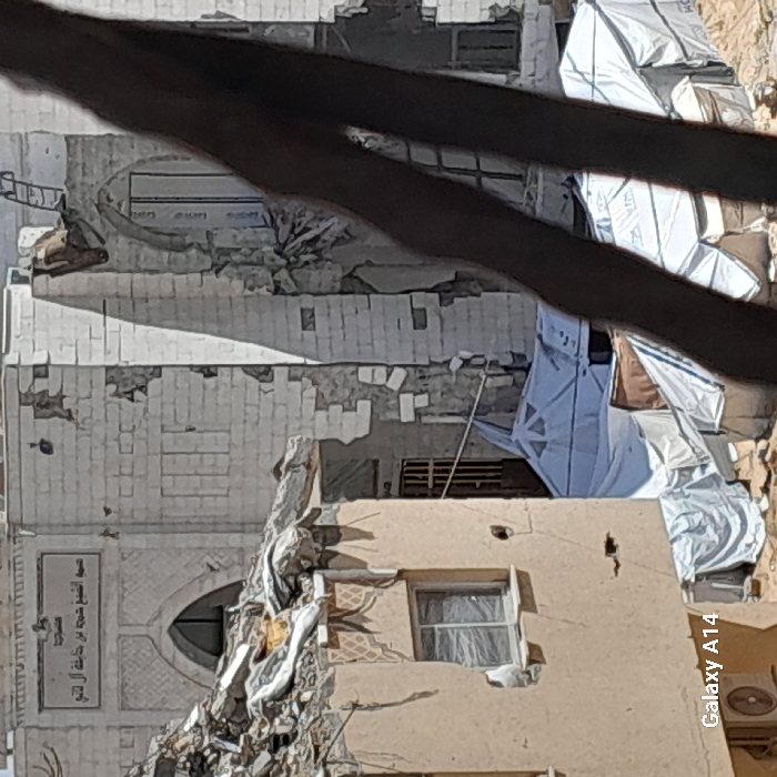 The image displays a severely damaged urban environment in Khan Yunis, Palestinian Territory, captured during daylight. In the left foreground, a multi-story building with a light-colored, possibly beige, facade shows a partially collapsed roof and significant structural damage, with rubble including twisted rebar and concrete resting on its upper section. A white-framed window is visible on an upper wall, and a partial view of an outdoor air conditioning unit is seen at the bottom left. In the midground, behind and to the right, a heavily damaged mosque built of light-colored stone or brick exhibits extensive destruction, with large sections of its walls missing or crumbled. A rectangular sign with Arabic script, translating to "Mosque of Sheikh Hamza," is mounted above an arched entryway. Further right, another tall, arched opening in the mosque appears boarded or sealed with white material. At ground level, amidst the debris and between the damaged structures, several white and some brown tarp-like temporary shelters or tents are erected. A metallic, barred gate is visible in the midground, and a ladder leans against a damaged wall in the upper right. A dark, out-of-focus object, likely a structural element or debris close to the camera, diagonally obscures the right portion of the scene. No individuals are visible within the frame.