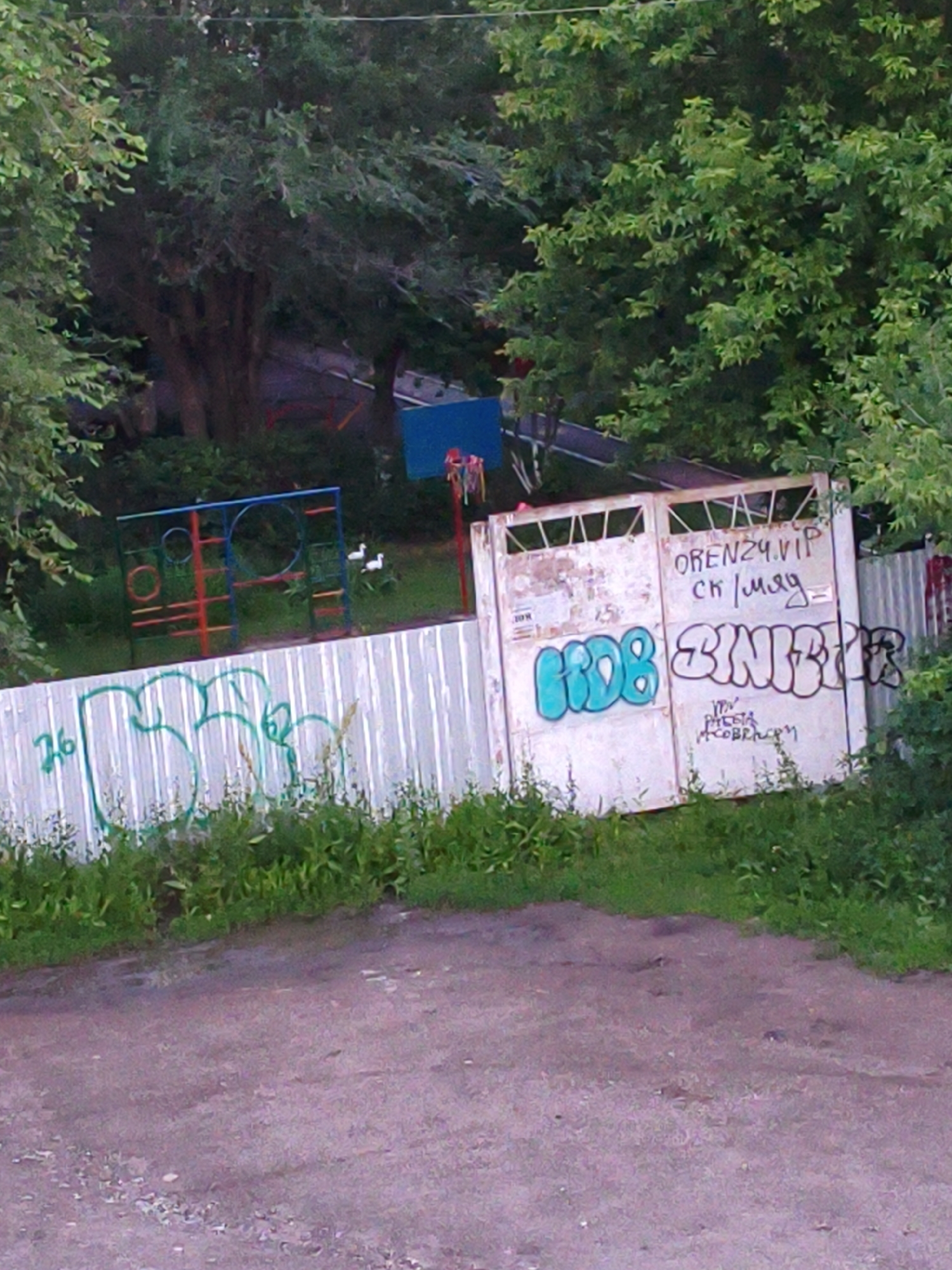 The image depicts an outdoor scene with a weathered white corrugated metal fence that has graffiti. To the left, the number "76" is spray-painted in green. To the right of "76" are large, stylized green graffiti letters that are difficult to decipher. Further to the right, on a white metal gate, there are two sections. The upper right section of the gate has black lettering that reads "OREN24.VIP SK (Illegible)". Below this, there is blue graffiti on the left section of the gate, spelling out "HOB", and white graffiti on the right section that appears to be an illegible tag. Beneath the white graffiti, there is smaller, illegible black text and a small drawing.
Behind the fence, a children's playground structure with red and blue metal bars is visible, along with some green grass and trees. Two white birds are in the background near the playground. The overall impression is an urban or semi-urban environment, likely an alleyway or a backyard area, given the presence of the fence, gate, graffiti, and playground equipment. The reference to "OREN24.VIP" strongly suggests the location is Orenburg, Russia. The ground in the foreground is a rough, reddish-brown dirt surface with some gravel. The lighting suggests it is daytime, possibly late afternoon, with shadows cast by the trees.