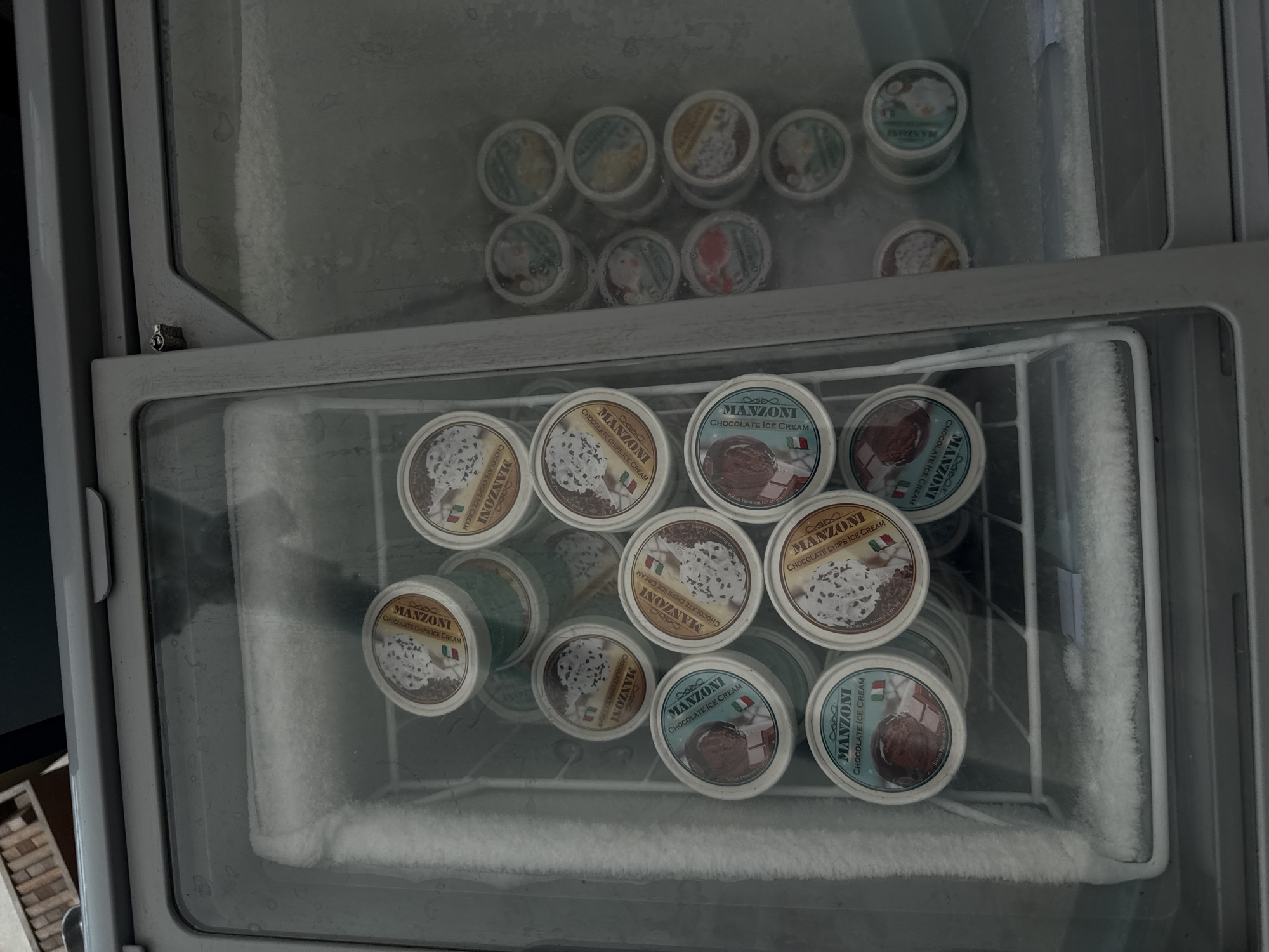 The photograph captures the interior of a freezer, presumably in a location within Rawai, Thailand, given the presence of "Manzoni" brand ice cream, an Italian premium ice cream company. The freezer is stocked with numerous single-serving containers of the ice cream, organized on the freezer shelves.
The upper shelf holds various ice cream flavors, while the lower shelf prominently displays several containers of "Manzoni" brand ice cream. The labels of the ice cream containers are visible, indicating flavors such as "Chocolate Chips Ice Cream" and "Chocolate Ice Cream." The presence of frost and ice crystals on the interior surfaces of the freezer and on some of the ice cream containers suggests the freezer is actively maintaining a low temperature. The freezer's wire rack shelves are also visible.