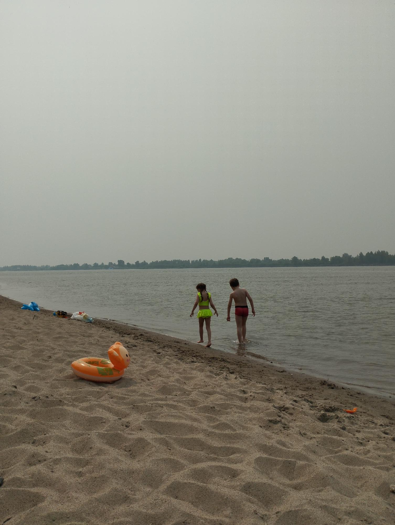 The image depicts a sandy beach along a wide river under an overcast sky. Two children are standing at the edge of the water, with one child, a girl wearing a neon yellow swimsuit, wading in the shallow water. Next to her, a boy in red swim trunks is also standing in the water. In the foreground, an orange inflatable toy shaped like a duck rests on the sand, and further up the beach, other toys and bags are visible. The background shows a treeline across the river. The scene is likely set in the vicinity of Bibikha, Russia, a village known for its river access.