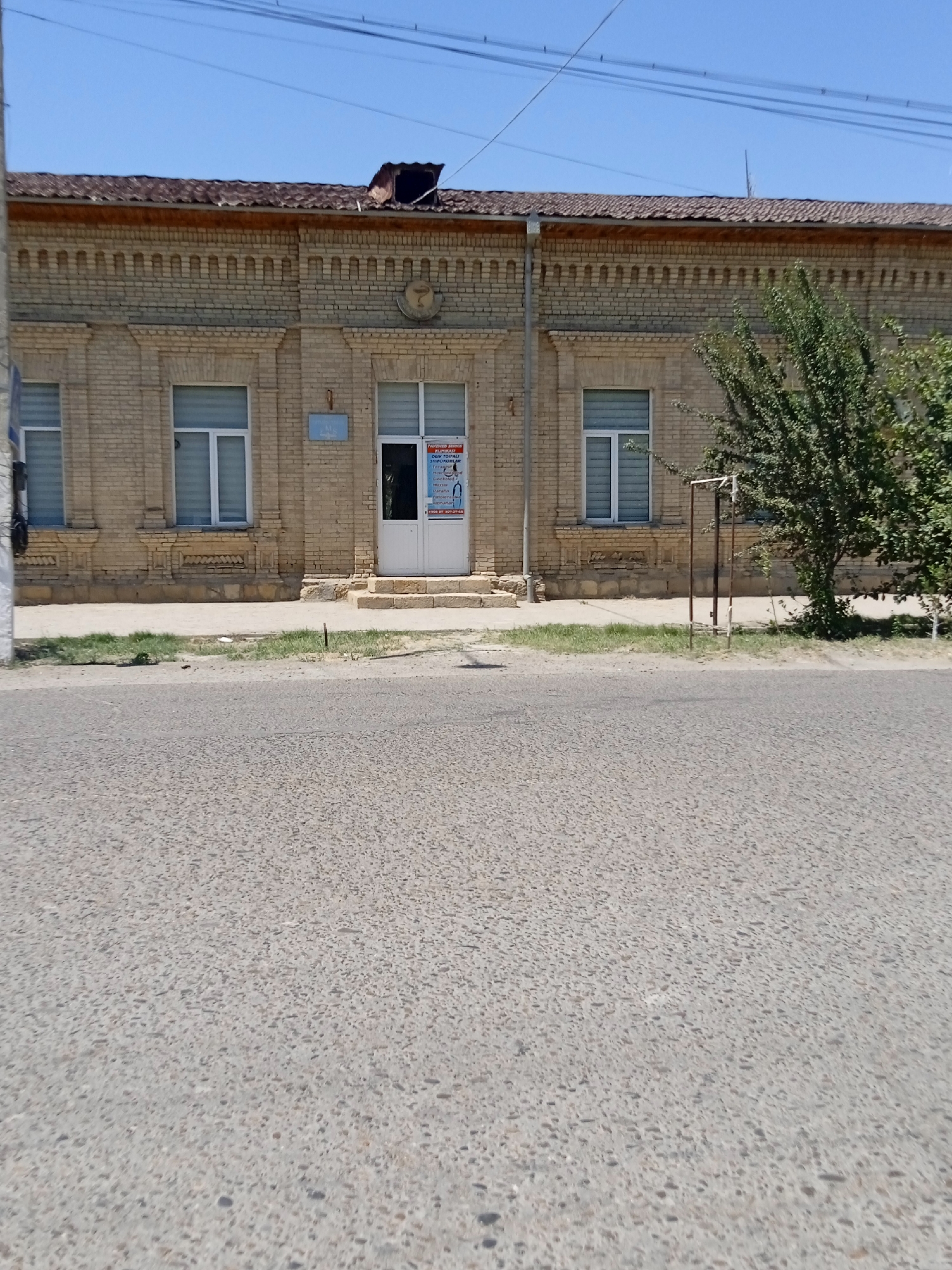 A frontal, eye-level, medium shot shows a light yellow brick building in the city of Unknown, Unknown. The building features decorative brickwork above the windows and a brown tile roof with a damaged section. The windows are rectangular with white frames and horizontal blinds. A white door is centered on the building, flanked by two sets of stone stairs. A sign with blue lettering is mounted above the door, and a poster with text and images is on the door itself. To the right of the building, a green tree with dense foliage is visible. The building is set back from a paved road, with a sidewalk and a patch of grass between the road and the building. Overhead power lines are visible against a clear blue sky.