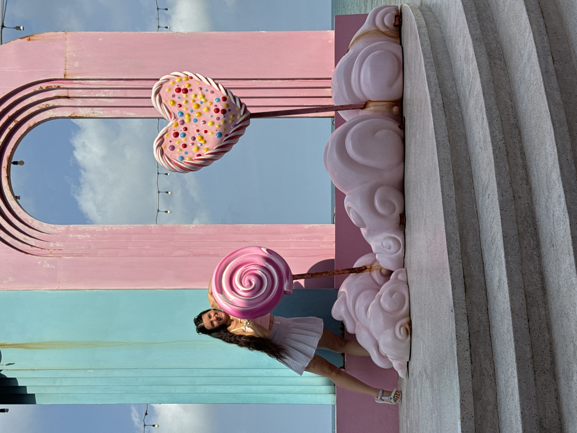 The image depicts a woman posing with two large, decorative lollipops in an outdoor setting. She is standing on a concrete platform with multiple steps, leaning against a pastel pink and turquoise structure that resembles an arched entryway. To her right, a large, heart-shaped lollipop with colorful candy dots is displayed, and to her left, a round, swirled lollipop is held by the woman. The lollipops are mounted on poles that emerge from pink cloud-like sculptures at the base. The background shows a clear blue sky with some clouds and the ocean in the distance. String lights are visible overhead. The location is identified as Tien Thanh Ward, Vietnam, suggesting this is a public art installation or decorative area.
