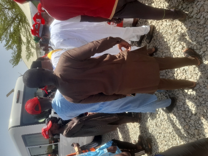 The scene is a street in Gusau, Nigeria, taken at 12.156 latitude and 6.709 longitude. A crowd of people are gathered in front of a white minivan. The person in the center of the image, wearing a brown shirt and pants, appears to be talking to another man wearing a white robe. The rest of the group is standing around, watching or conversing. All of the individuals are wearing clothing typical of the region, and some are wearing caps with red lettering. The terrain is composed of loose gravel. The scene is brightly lit, with a clear, blue sky in the background.