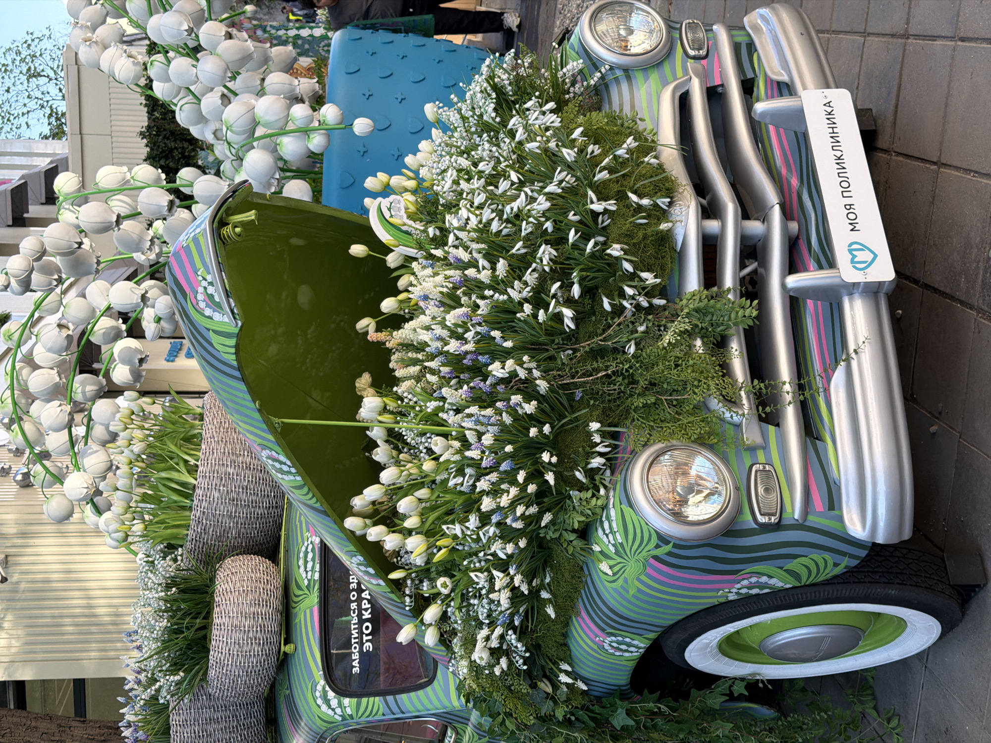 The image depicts a vintage car, adorned with flowers, in an outdoor setting that could be interpreted as an urban display or art installation. The car itself is painted in a decorative, wavy green and grey pattern. It has a license plate on the front bumper with a blue logo and Cyrillic text "Моя поликлиника", which translates to "My polyclinic" (My clinic) suggesting a connection to a healthcare facility or initiative. The car is heavily decorated with white and purple flowers, and greenery, filling the hood and spilling out from the interior. The tires have whitewalls, and the wheels are painted green. There are wicker baskets placed on the rear. In the background, there are large lily-of-the-valley-like decorations. Given the Cyrillic script on the license plate and overall style, it's plausible that this scene is in Moscow, Russia.