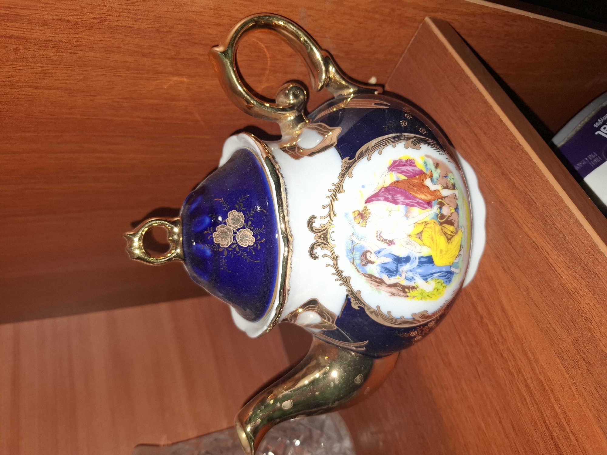 A porcelain teapot with a blue lid adorned with gold floral patterns sits on a wooden shelf. The teapot is decorated with a scene depicting two women in classical attire, one in a blue gown and the other in a yellow robe, interacting with a vase. The teapot has a gold spout and handle. The background is a wooden surface with visible grain. The image appears to be taken indoors, possibly in Almaty, Kazakhstan, though no specific landmarks are visible to confirm this location.