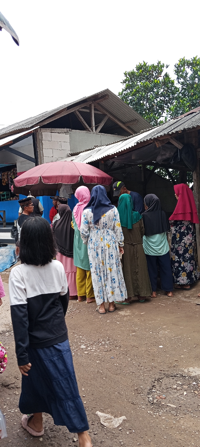 The image shows a group of people standing outside a small, open-sided store. There are 8 people in total, including a young girl with black hair in a white and black sweater and a long blue skirt who is standing near the bottom of the frame. The girl is facing slightly to the right, and her left leg is extended forward, while her right leg is bent at the knee. Her left foot is resting on the ground, while her right foot is off the ground.
The people are standing in a line, facing the store. The woman in the front of the line is wearing a white floral dress and has a pink scarf on her head. The woman behind her is wearing a green dress. The other people in the line are wearing various clothing, including headscarves and traditional clothing.
The store has a corrugated metal roof and wooden support beams. There is a red umbrella on the left side of the store. The image is likely taken in Kecamatan Kutawaringin, Indonesia, based on the coordinates provided. The ground is rough and dusty, and there are some pieces of trash on the ground. The scene is typical of a bustling, everyday market scene in Indonesia.