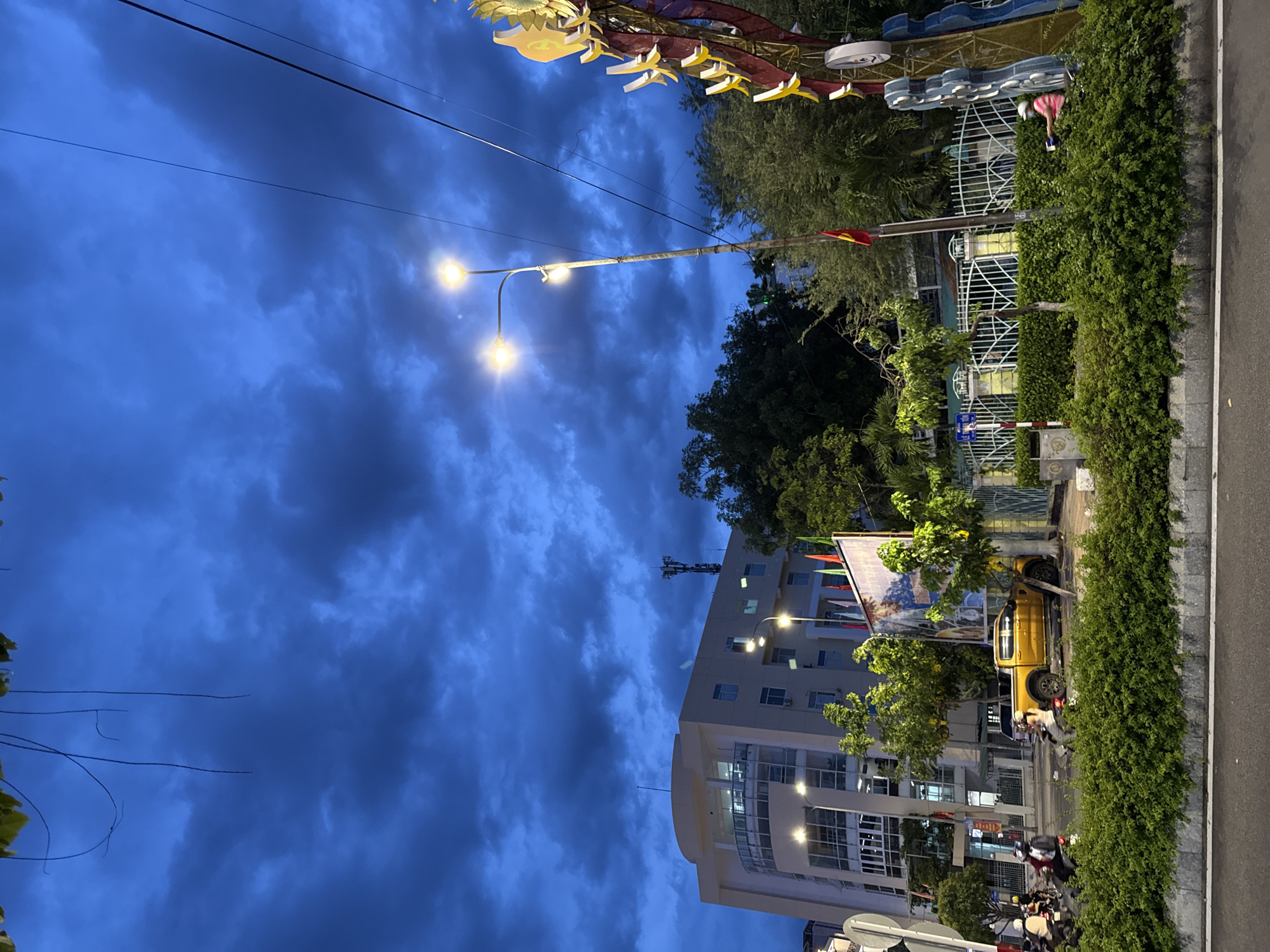 The image depicts a street scene in Nha Trang, Vietnam, at dusk. The sky is a deep blue with dark, dramatic clouds. On the left side of the image, a modern, multi-story building with rounded corners and large windows is visible. Streetlights illuminate the area in front of the building. A bright yellow pickup truck is parked on the side of the road, partially obscured by greenery. Several people are seen on motorbikes, some appearing to be riding on the road and others parked. A dense hedge runs along the foreground, separating the road from a sidewalk or walkway. In the background, trees and foliage are visible, as well as a metal fence and a large, decorative structure on the right side of the frame, featuring yellow and red elements. A Vietnamese flag is planted on a pole near the fence. The overall impression is one of an urban environment at the end of the day, with signs of activity and infrastructure.