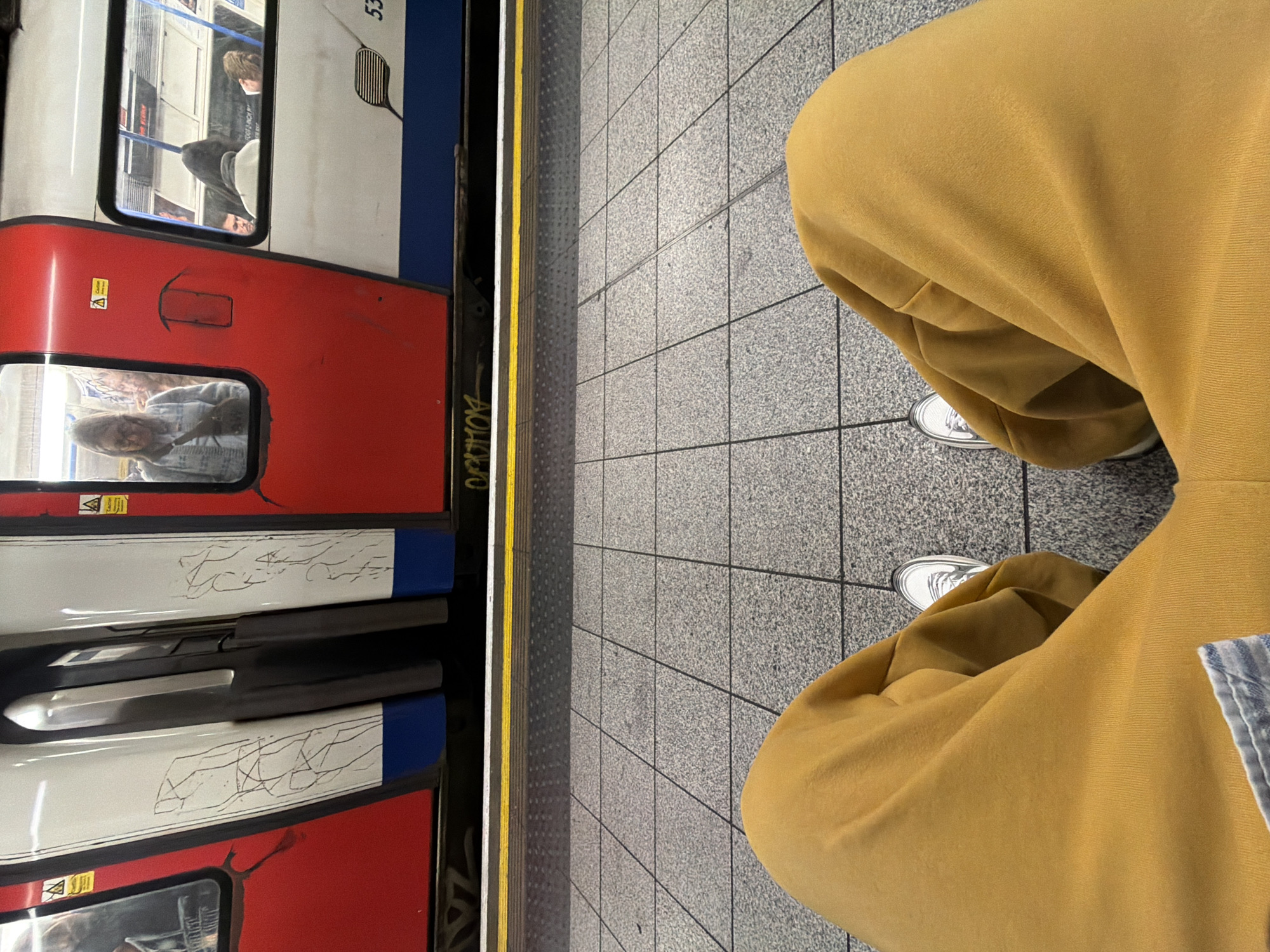 An eye-level, ground-up shot captures a person wearing a mustard-yellow tracksuit and white trainers, standing at a London Underground station. The edge of a tube train is visible, with red and white carriages and blue accents. Passengers can be seen through the train windows. The station platform is made of grey square tiles, demarcated by a textured yellow safety line.