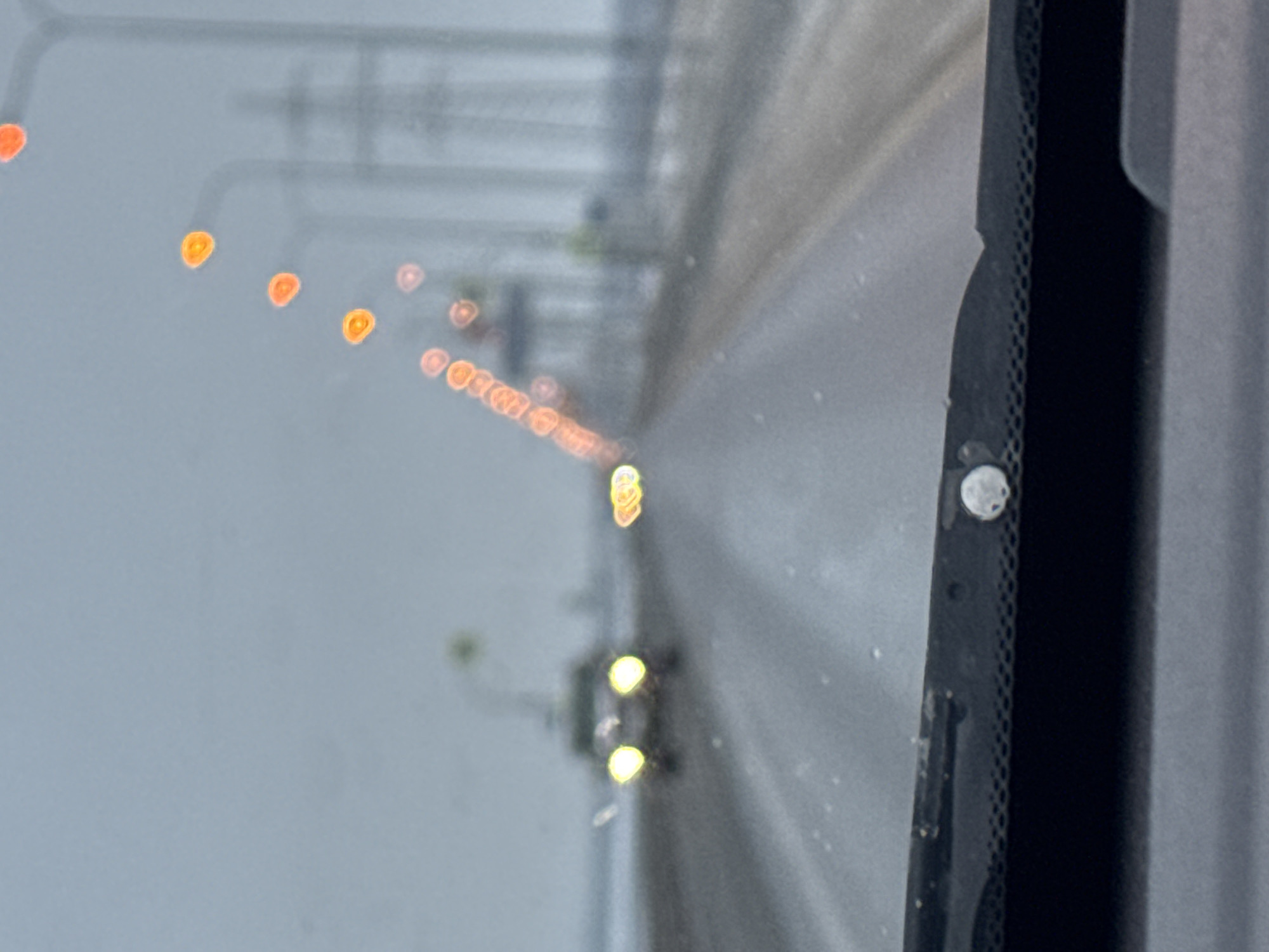 The image shows a view through a car windshield on a road, likely a highway, in Noviy, Russia. The road is mostly obscured by blur but appears to be grey and wet. The lights of oncoming traffic appear as blurred orange and yellow orbs in the distance. Utility poles or power lines are visible in the background, also out of focus. The windshield wiper mechanism is visible in the foreground, attached to a grey frame, obscuring the view on the right side.