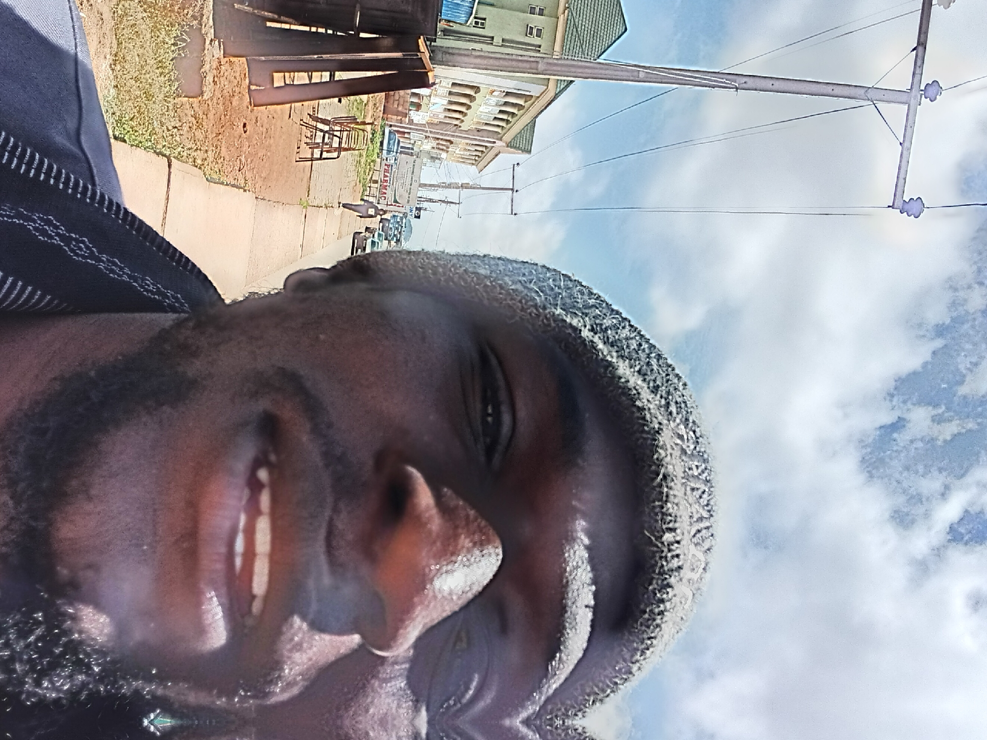 A close-up selfie shows a man wearing a knitted beanie and a dark blue garment with a patterned neckline. He is outdoors, with a street scene visible in the background. The background features buildings with green roofs and arched windows, a sidewalk, a road with vehicles, and utility poles with wires. The sky is partly cloudy, and the lighting suggests it is daytime. The man's face is the primary subject, with his eyes, nose, and mouth visible. The image is slightly distorted due to the wide-angle lens and close proximity to the subject.