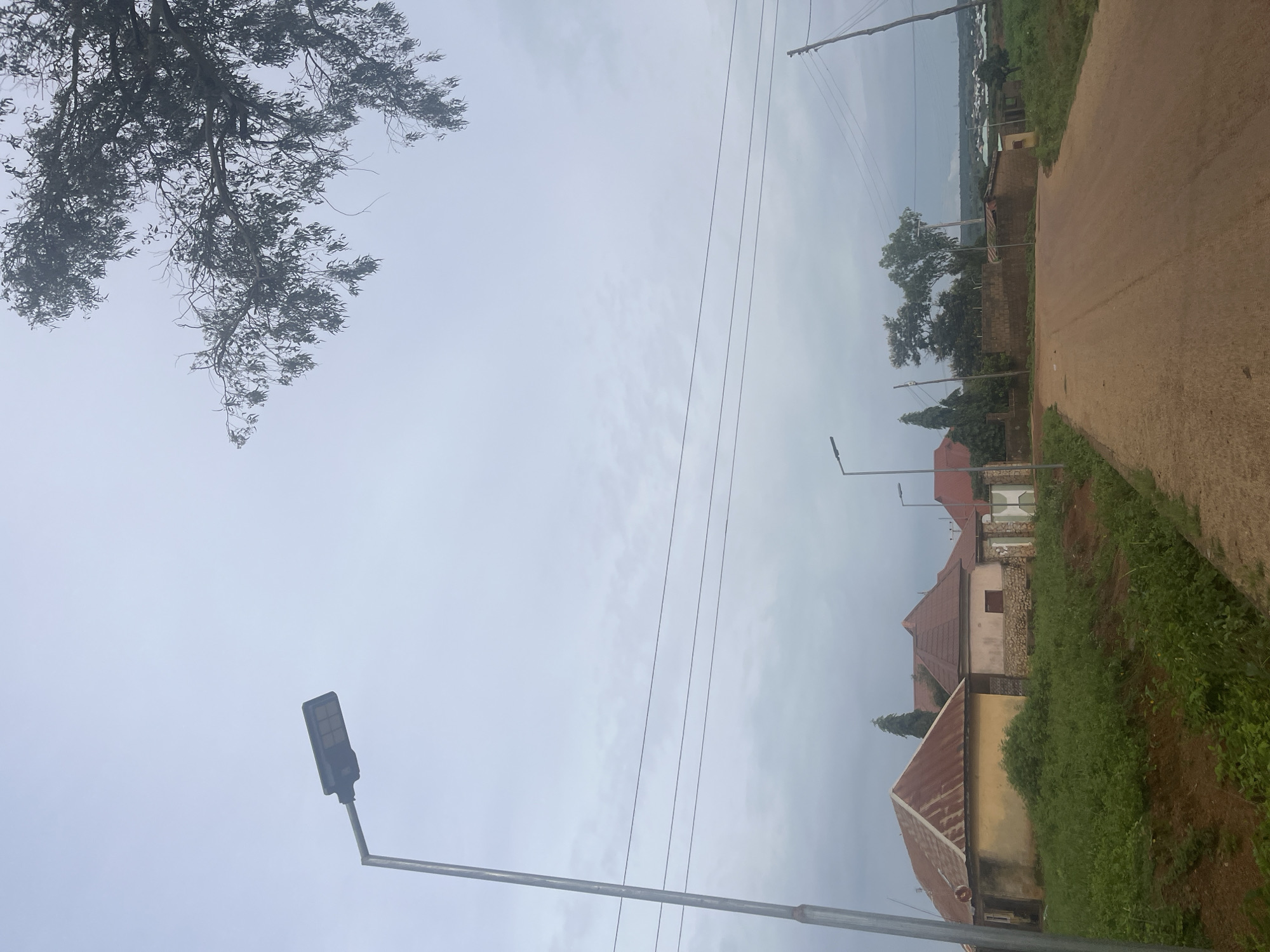 The image depicts a dirt road in Unguwar Saya, Nigeria. A tall streetlamp is positioned on the left side of the road. Several houses with red corrugated metal roofs are visible on the left, some with yellow or stone-colored walls. Further down the road, more houses and trees can be seen, with a cluster of buildings and vegetation in the distance. Power lines stretch across the sky, supported by poles. A tree with dense foliage hangs over the scene from the upper right corner. The sky is overcast.
