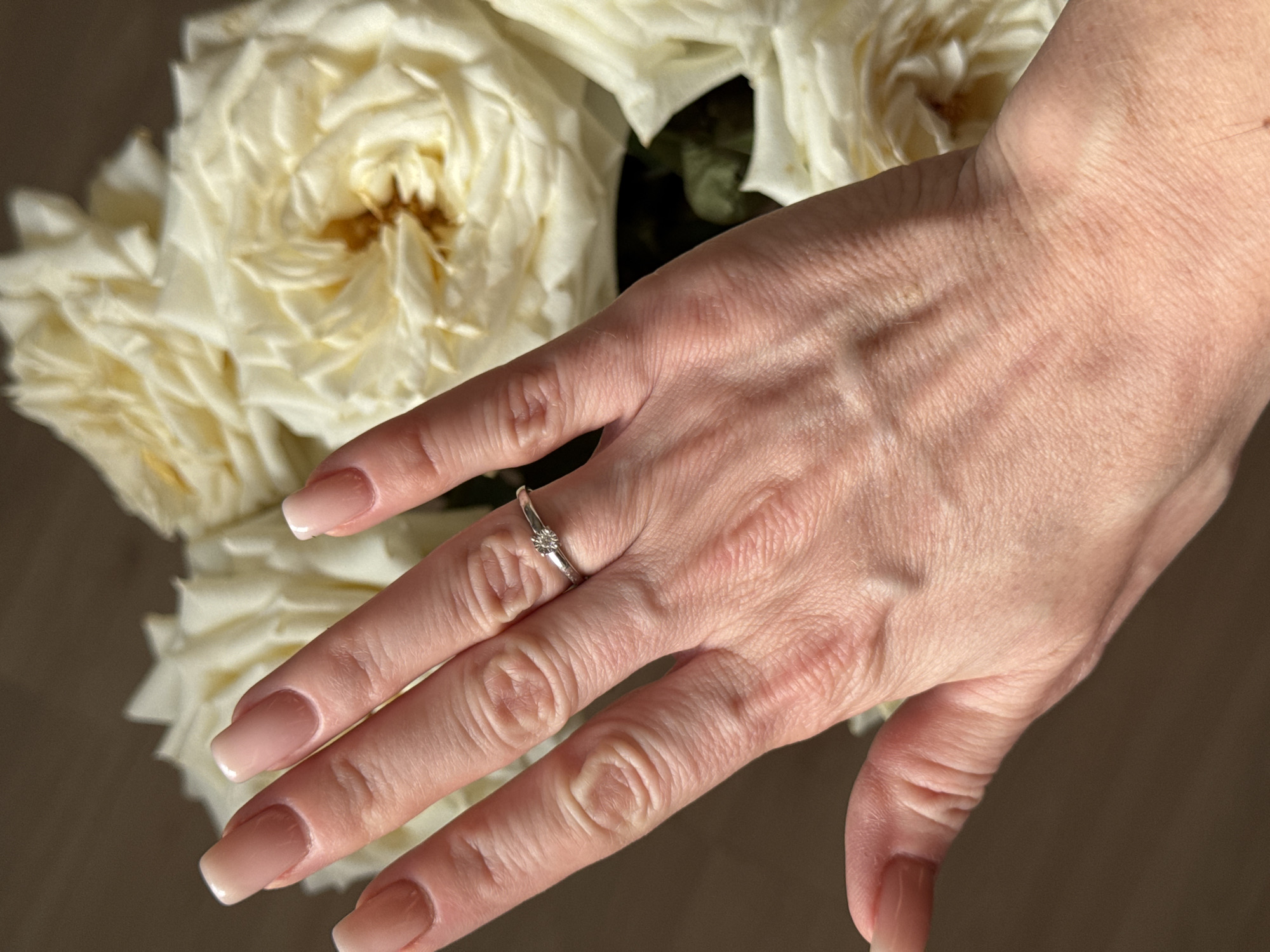 A woman's hand, adorned with a simple engagement ring on the ring finger, is positioned against a backdrop of white roses. The hand's skin shows visible wrinkles and veins. The nails are manicured with a neutral color and a classic French tip. The scene is dimly lit. The image provides no information to place the scene in Kazan, Russia or any other city.