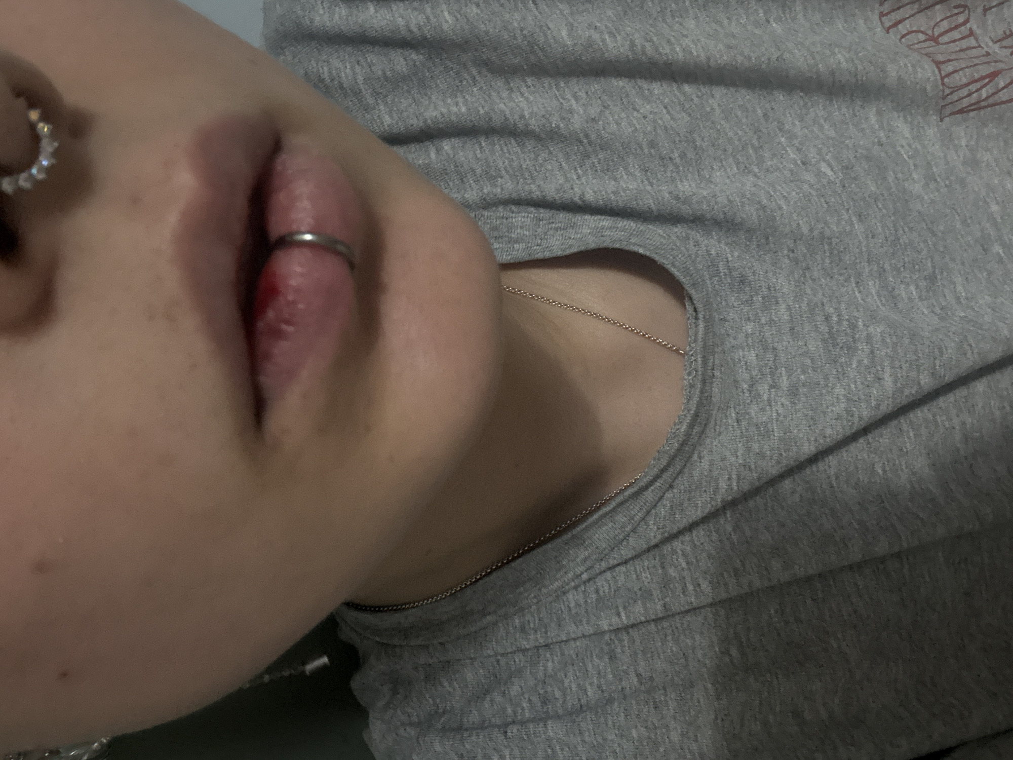 An angled close-up shows a person from the nose down to the upper chest. A nostril is pierced with a ring of stones. The lower lip is pierced with a silver ring, and some blood is visible. The person wears a gray t-shirt and a delicate silver chain around their neck. The background is blurry and out of focus. No information suggests this image was taken in Saint Petersburg, Russia.