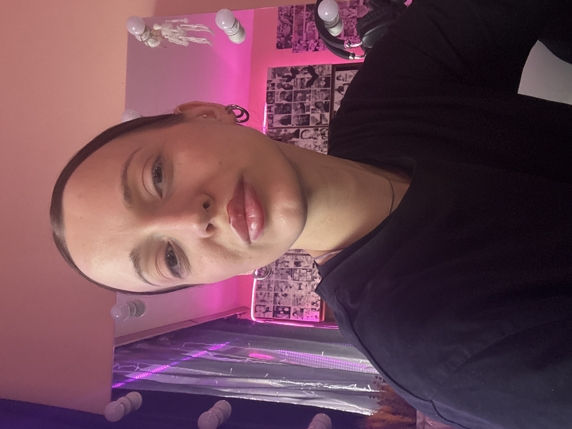A close-up shot of a fair-skinned person with dark hair pulled back, positioned at an angle. The person's face is turned toward the camera, featuring full lips and visible eye makeup. They are wearing a dark shirt. The background includes a mirror surrounded by lights, pink walls, and various decorations including what appears to be manga panels and a small skeleton figure. A ring light with a microphone attached is partially visible in the upper right. The location is not discernible from the image.