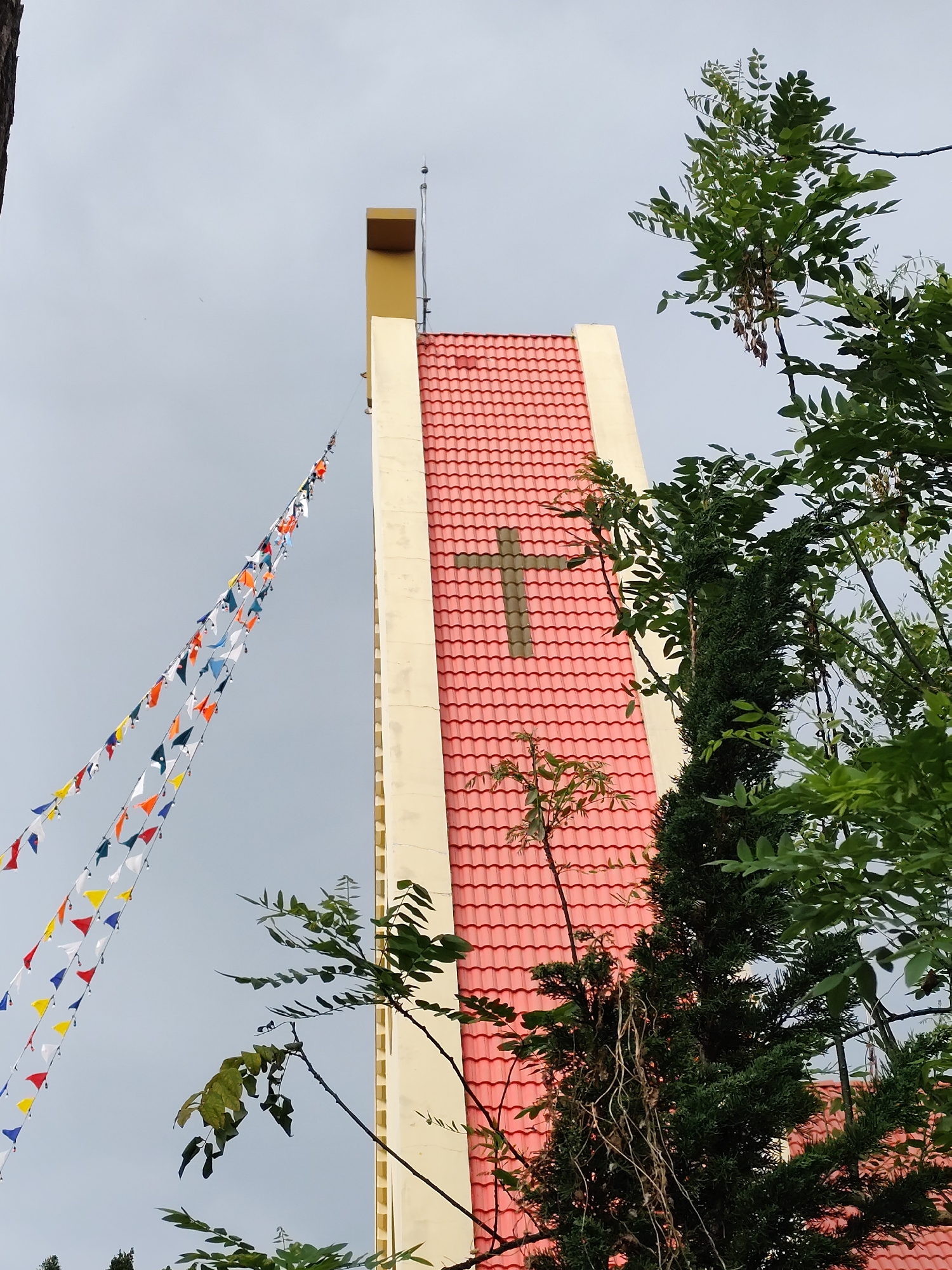 A low-angle shot shows a church steeple and trees against a cloudy sky. The steeple is a rectangular tower with a cross-shaped shadow in the center. The tower is a pale yellow on either side and light red brick in the middle. The top of the tower is capped with yellow and a metal rod. Stringed flags in various colors hang to the left of the tower. Green trees partially obscure the right side of the tower. This location is in Da Lat, Vietnam.