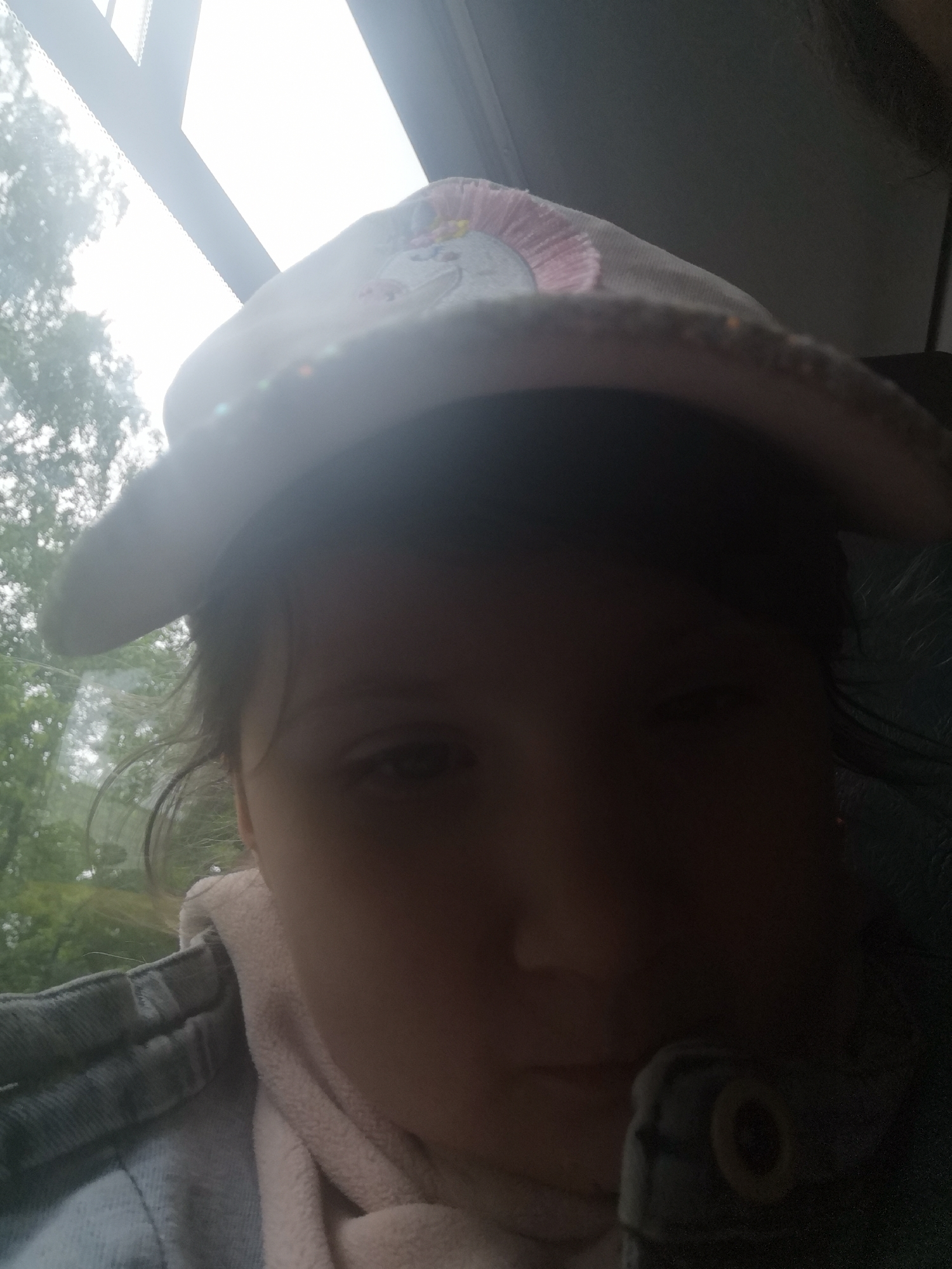 The image is a back-lit portrait of a young person, likely a child, taken inside a vehicle. The subject is positioned on the left, wearing a light-colored hat with decorative stitching or sequins, a denim jacket, and a light-colored, soft-textured top or scarf. Their face is heavily shadowed by strong light coming from a large window on the left, but one eye is partially visible, showing a neutral or pensive expression.
Outside the vehicle's window, green trees and foliage are visible against a very bright, possibly overcast or rainy daytime sky. The window frame is distinct. The deep shadows within the vehicle's interior obscure most details, though a faint outline of another person's head or hair can be seen on the far right. The overall impression is of a journey, possibly in a car, bus, or train. The location context is Chaikovsky, Russia. No text is visible in the image.
