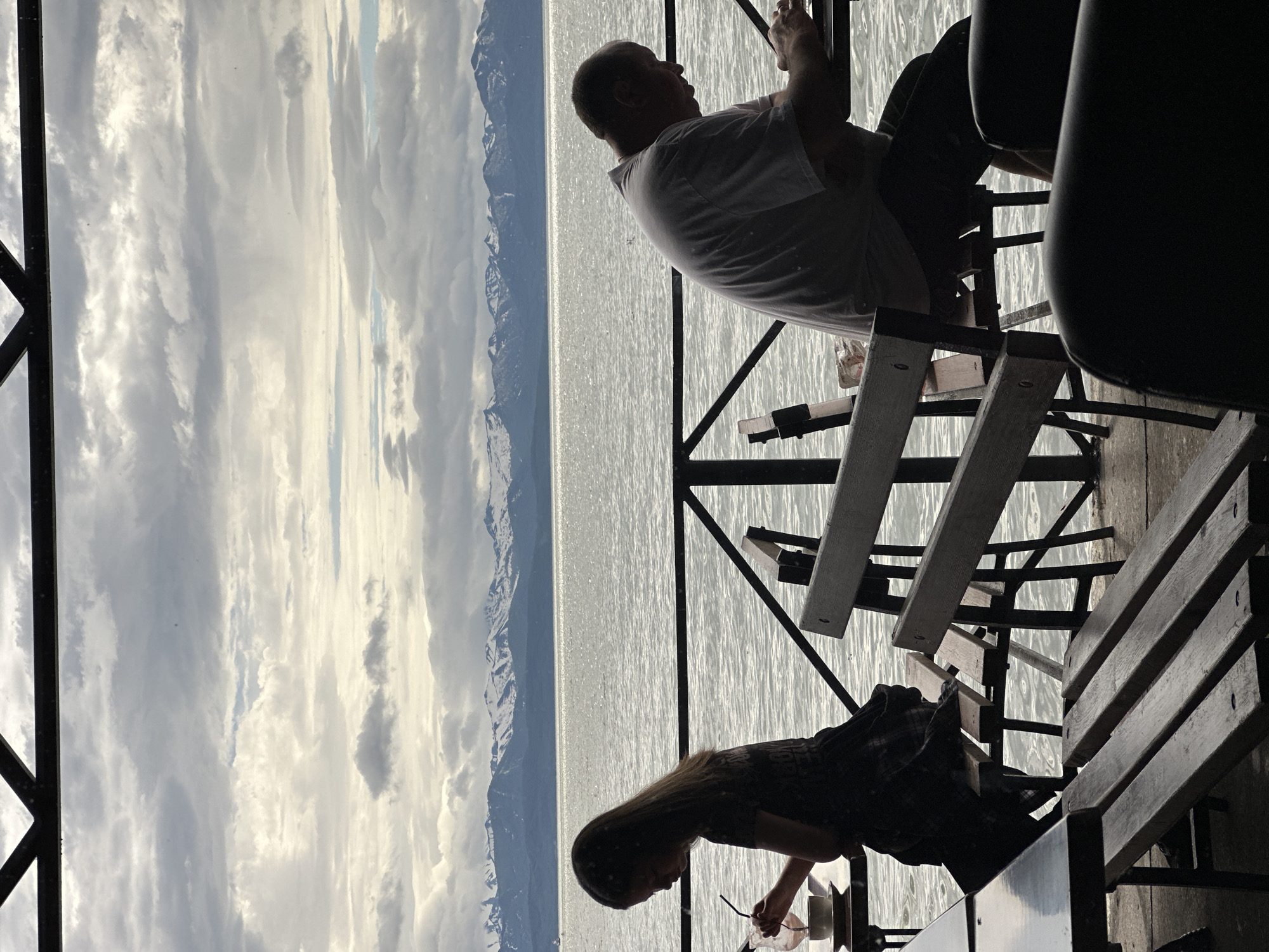 The image depicts an outdoor seating area overlooking a body of water, with snow-capped mountains in the background. Two individuals are seated at separate tables. On the left, a young person with long hair is sitting at a table, holding a straw in a drink and looking down. On the right, an adult male is seated at another table, looking down and appearing to use a laptop or tablet. Both individuals are silhouetted against the bright, overcast sky and the glistening water. Wooden benches and tables are visible in the foreground, and a metal railing separates the seating area from the water. The scene suggests a potentially casual dining or relaxation spot with a scenic view. The presence of snow-capped mountains suggests a location in a colder climate or at a higher altitude, possibly Petropavlovsk-Kamchatskiy, Russia, which is known for its stunning natural landscapes including volcanoes and mountains.
