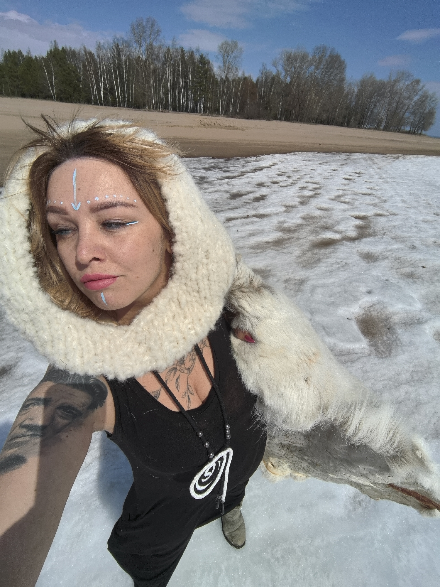A medium shot features a person, likely a woman, in an outdoor, snow-covered landscape, possibly in Noviy, Russia. She is taking a high-angle selfie, looking downwards with a pensive expression.
The subject has light brown, slightly disheveled hair and distinctive face paint, including a light blue arrow pointing down on her forehead, small white dots above her eyebrows, and light blue lines extending from her right eye and below her lower lip. She wears pink lipstick. Her attire consists of a black tank top or dress, a large, chunky white knitted hood, and a white fur stole draped over her left shoulder and arm. A necklace with a prominent white spiraled pendant and dark beads adorns her neck. Visible tattoos include a large portrait on her right arm and a floral design on her chest. Light-colored boots are visible on her right foot.
The scene depicts a natural environment with patches of melting snow mixed with areas of bare brown earth or sand. A sandy bank rises in the background, topped by a line of trees; some are deciduous with bare, light-colored trunks, while others appear to be evergreens. The sky above is clear and blue with minimal clouds. The image captures a sunny day during late winter or early spring in a natural, somewhat rugged setting. No visible text is present.