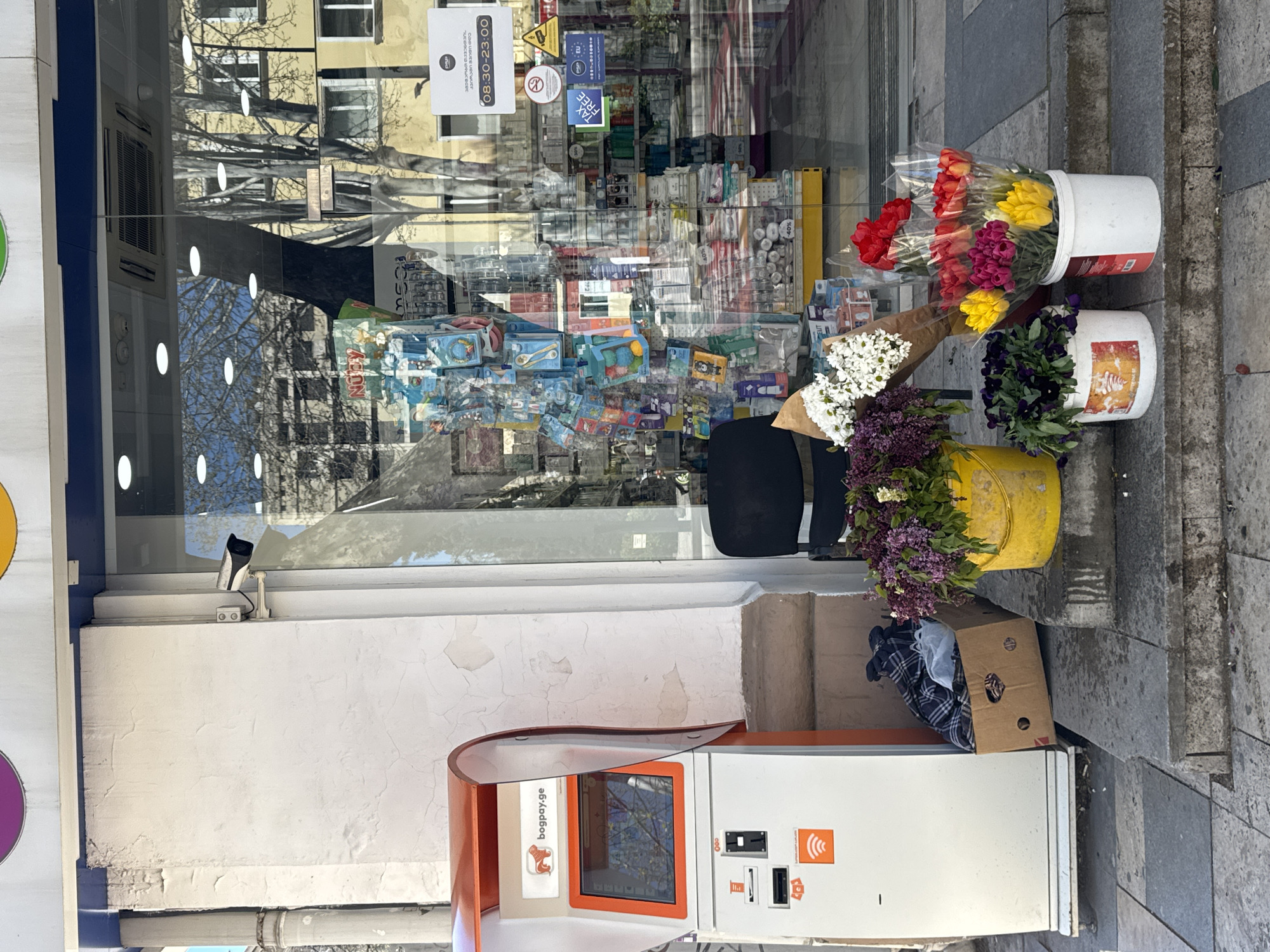 A street scene in Tbilisi, Georgia shows an ATM-style payment kiosk next to a storefront. Buckets containing cut flowers are placed on the sidewalk. The storefront's glass reflects the city environment.