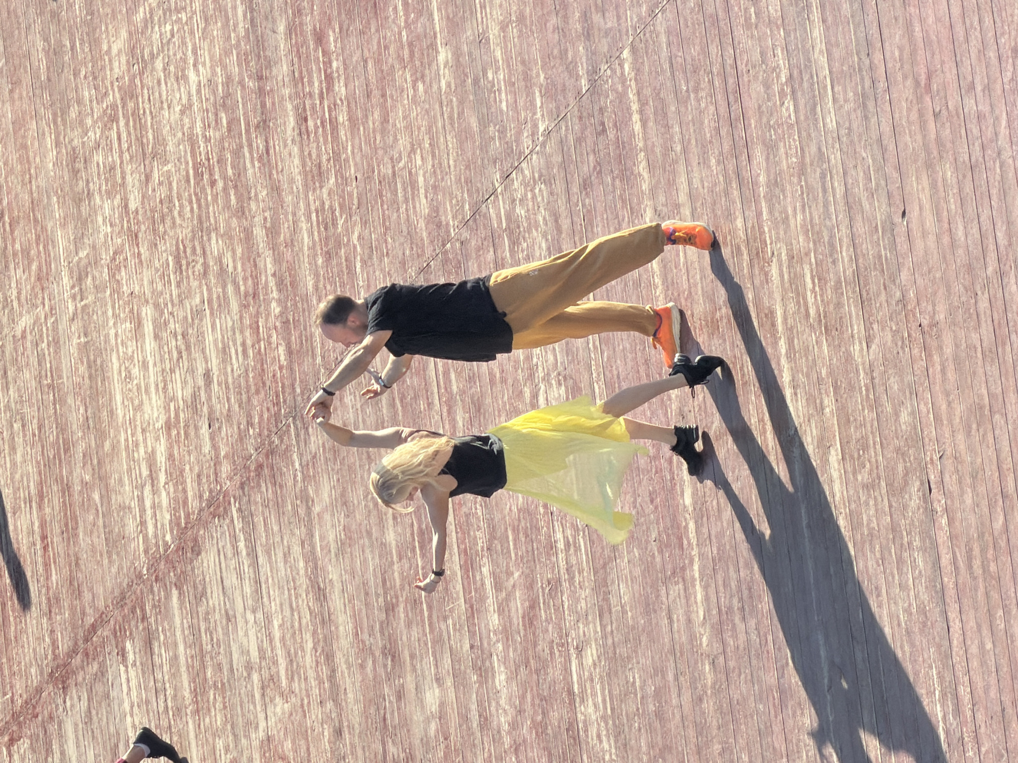 An overhead shot depicts a man and a woman in a plank position, holding hands on a reddish-brown, vertically-lined wooden surface. The man wears a black shirt, tan pants, and orange sneakers, and the woman is dressed in a black top, yellow skirt, and black boots. The location is Moscow, Russia.