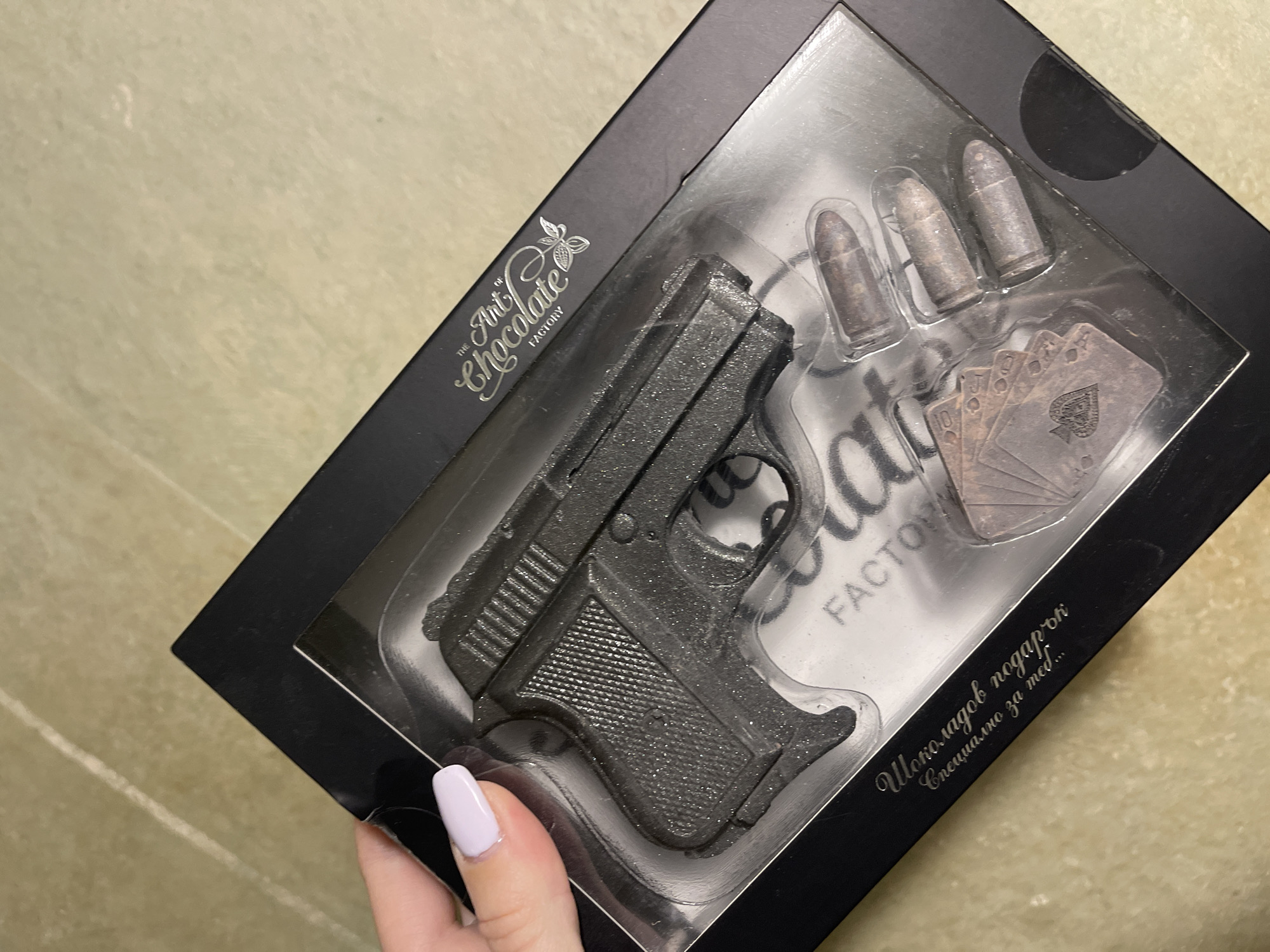 The image shows a black box containing chocolate novelty items. Inside the box, visible through clear plastic packaging, are chocolate representations of a handgun, three bullets, and a hand of playing cards with the Ace of Spades showing. The box is labeled "The Art Chocolate Factory." There is also Cyrillic text in the corner of the box reading "Шоколадов подарък специално за теб..." (Chocolate gift, specially for you...). The box is held by a hand with lavender nail polish. The background is a neutral beige color. The location is likely Sofia, Bulgaria, based on the Cyrillic text.