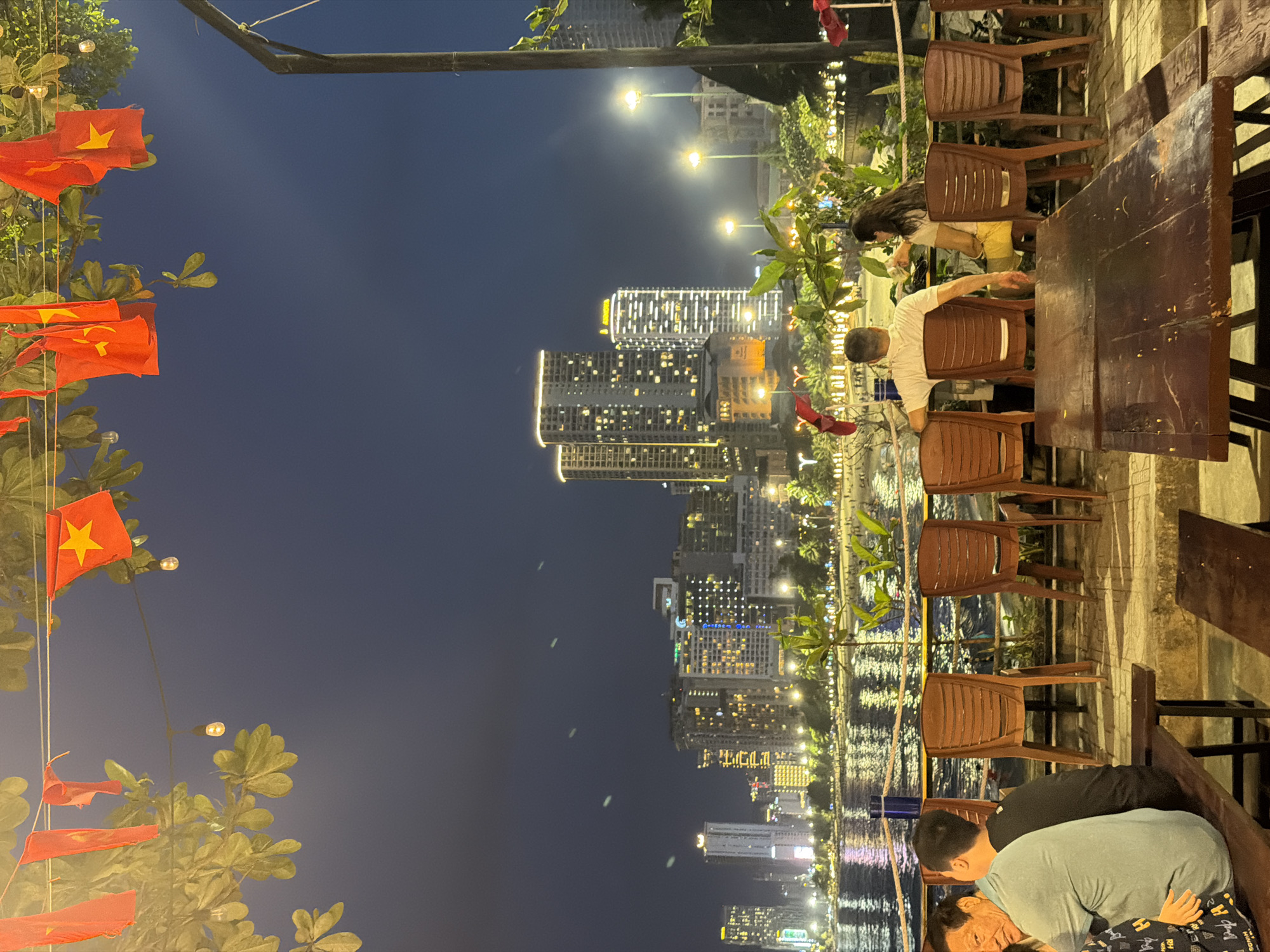 In Nha Trang Ward, Vietnam, a nighttime scene unfolds at a waterfront establishment. In the foreground, patrons sit on benches and chairs, facing away from the camera towards a body of water reflecting the lights of a city skyline. Vietnamese flags are strung overhead, interspersed with lightbulbs, against a dark sky. The city skyline features numerous illuminated high-rise buildings. The environment appears to be an outdoor seating area overlooking a beach and the ocean.