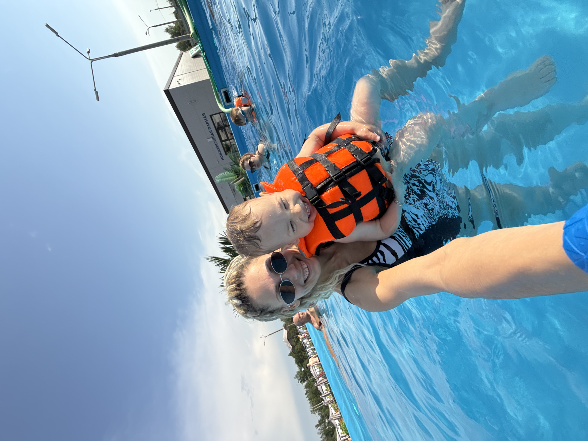 The image shows a woman holding a young child, both in a swimming pool. The child is wearing an orange life vest. The woman has sunglasses on and is smiling. The pool is outdoor, with other people visible in the background. In the background, there is a building with text in Russian and some vegetation. Street lights are visible. It is daytime, with a blue sky. The location is in Rusanovo, Russia.