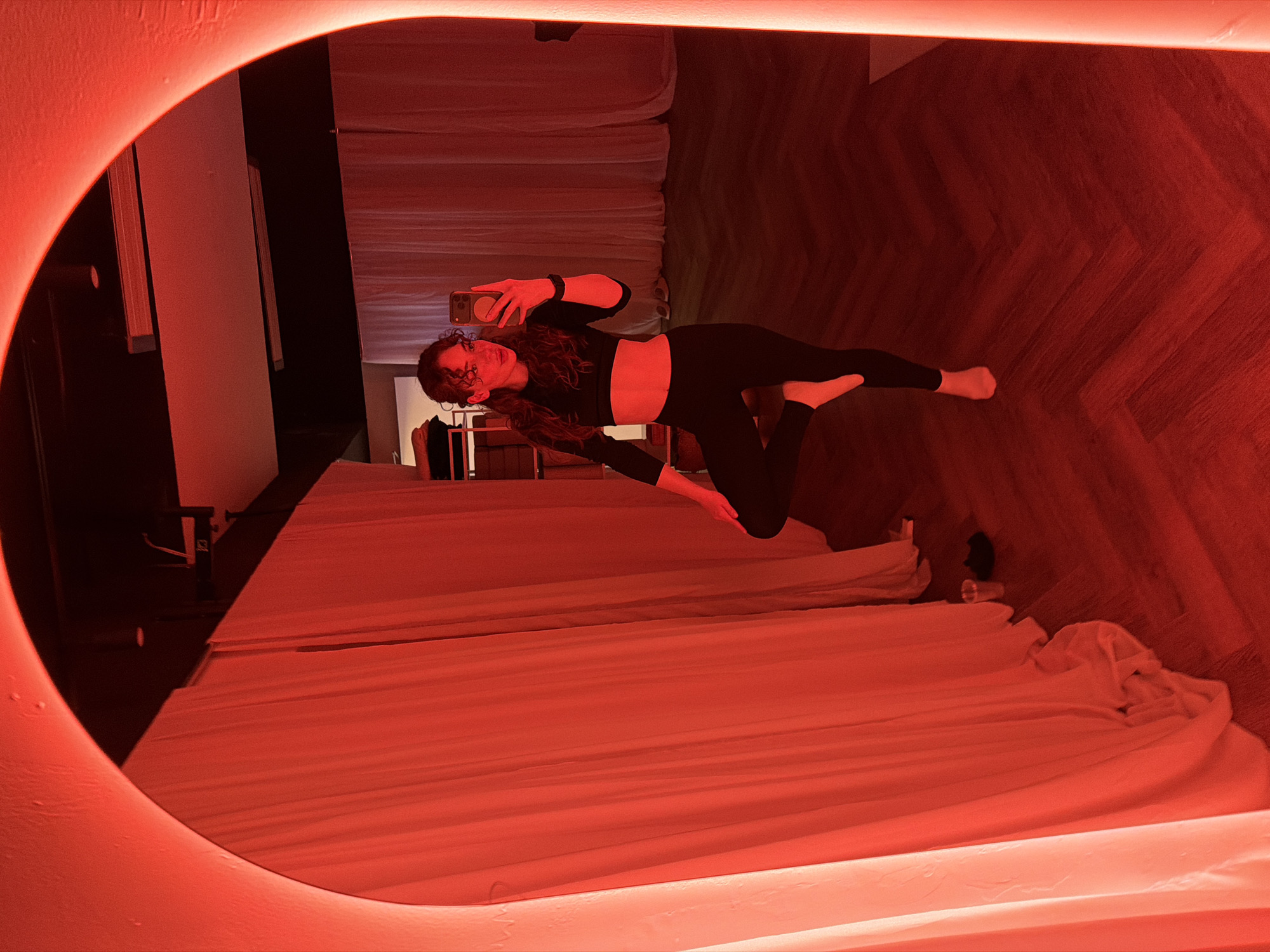 A woman in athletic attire is seen taking a mirror selfie inside a room illuminated with red lighting. She's wearing black leggings and a black crop top and is barefoot. The reflection shows she is holding a smartphone in her hand. The room features a large bed with white sheets, a wooden floor, and a dark-colored wall. The mirror itself has a rounded shape with built-in red lighting around the edge. The location is not Moscow, Russia.