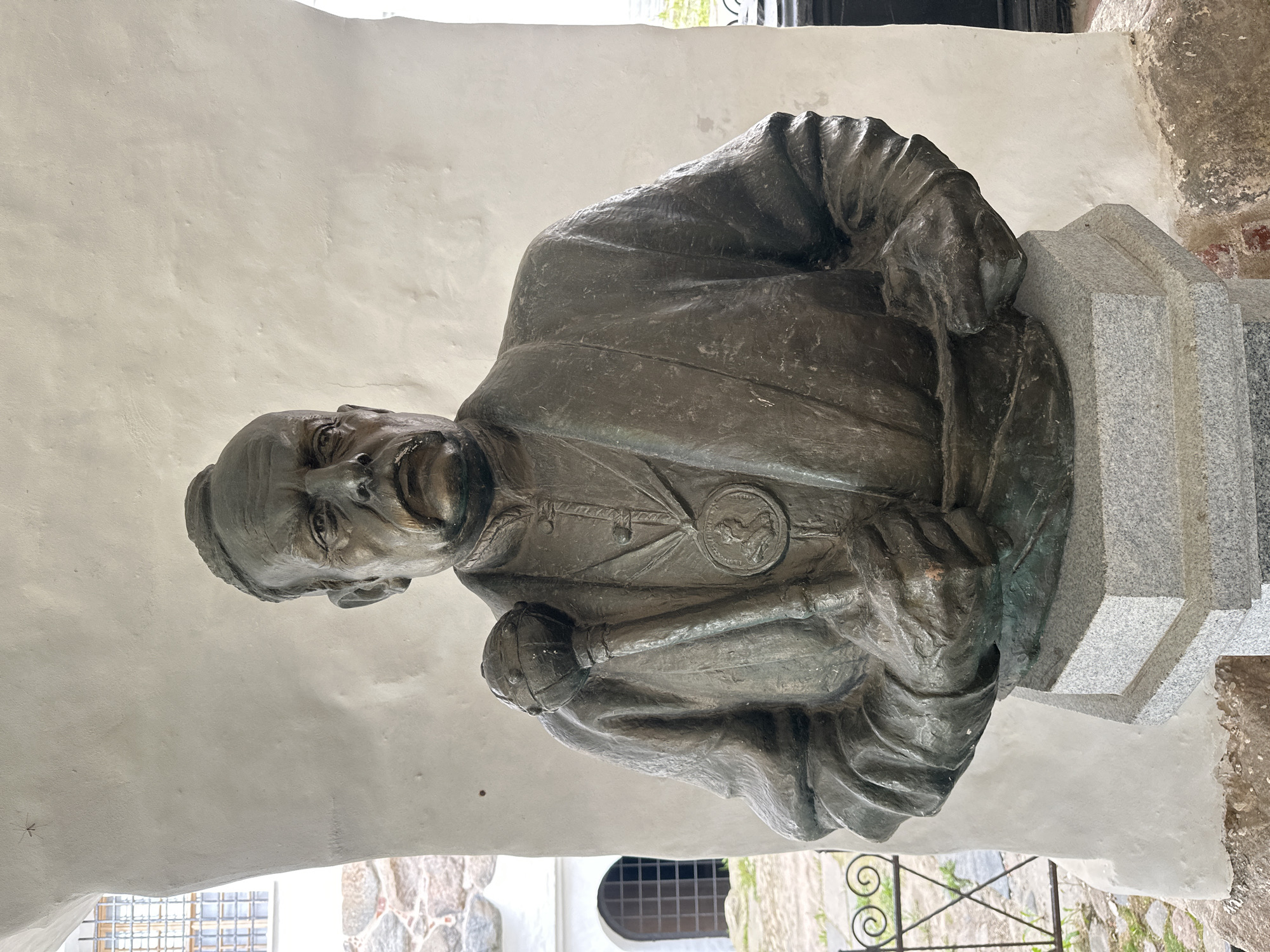 A bronze bust of a man with a mustache stands on a granite pedestal. The man is wearing a jacket with a medal around his neck and is holding a mace in his right hand. The bust is located in Solovetskiy, Russia, specifically on the grounds of the Solovetsky Monastery. The background shows a white stucco wall with arched windows and stone buildings.
