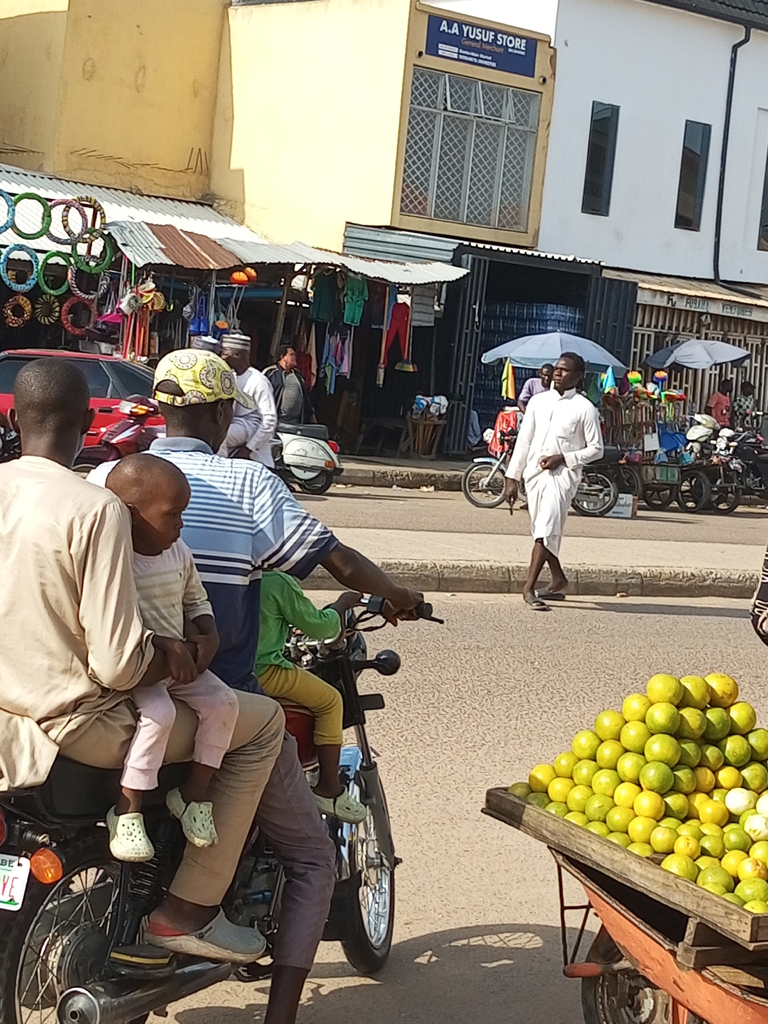 The image depicts a street scene in Arawa, Nigeria, with a focus on a group of people and their modes of transport. In the foreground, two individuals are seated on a motorcycle. The person in front is wearing a light-colored traditional garment, and is accompanied by a young child on their lap. The child is wearing light-colored trousers and a striped shirt. Behind them, another person in a blue and white striped shirt is also on the motorcycle, wearing a patterned cap. They are in motion, suggesting they are traveling.
In the background, several other individuals are visible. A man in a white traditional outfit is walking across the street. Other people appear to be standing near shops or stalls lining the street. Various objects are present, including motorcycles, a cart laden with yellow fruits, and stalls displaying goods such as clothing and what appear to be colorful circular decorations. The buildings in the background are simple, with some having awnings and storefronts. The overall environment appears to be an urban commercial area, with evidence of daily life and commerce.