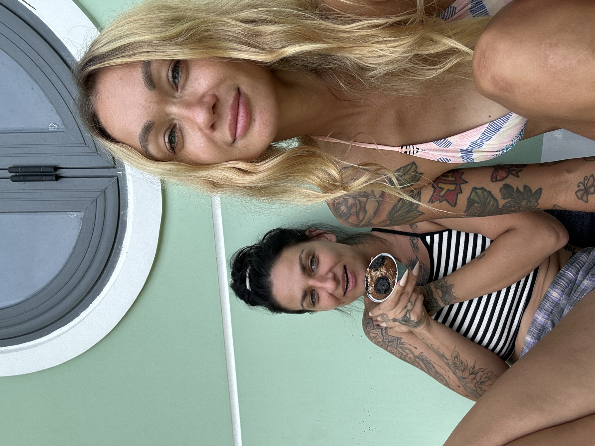 Two women are sitting outdoors, with a pale green wall behind them and a curved window with a dark grey frame in the upper portion of the image. The woman on the left has dark hair pulled up and is wearing a black and white striped top and plaid shorts. She is holding a muffin in her right hand, which is near her face. She has many tattoos on her arms and chest. The woman on the right has long blonde hair and is wearing a pink and blue striped bikini top. She has tattoos on her left arm and shoulder. Both women are looking at the camera. The setting appears to be a residential area or a cafe in Mũi Né, Vietnam.